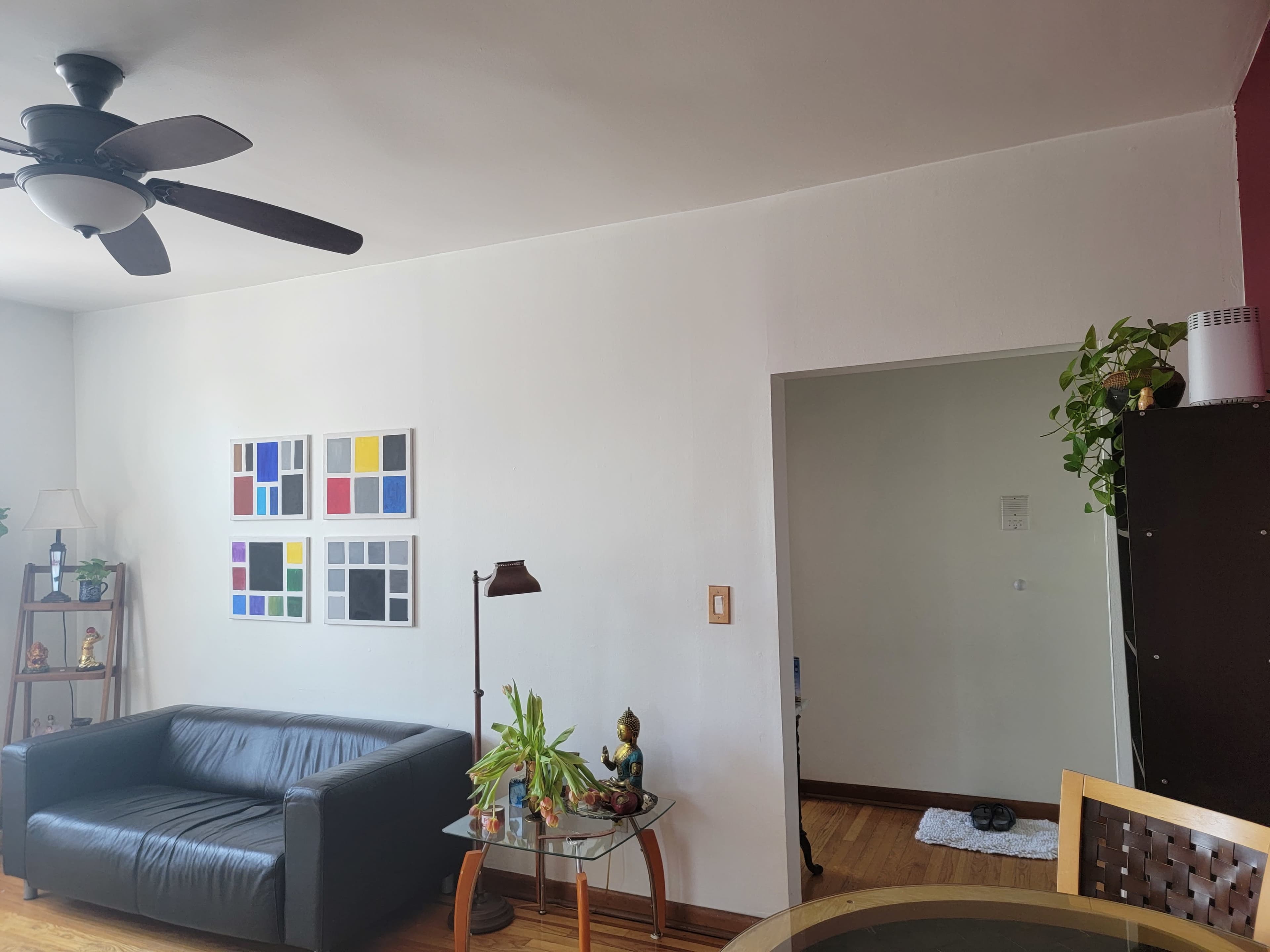A living room with a black couch, framed colorful artwork on the wall, a standing lamp, and a doorway leading to another room with greenery.