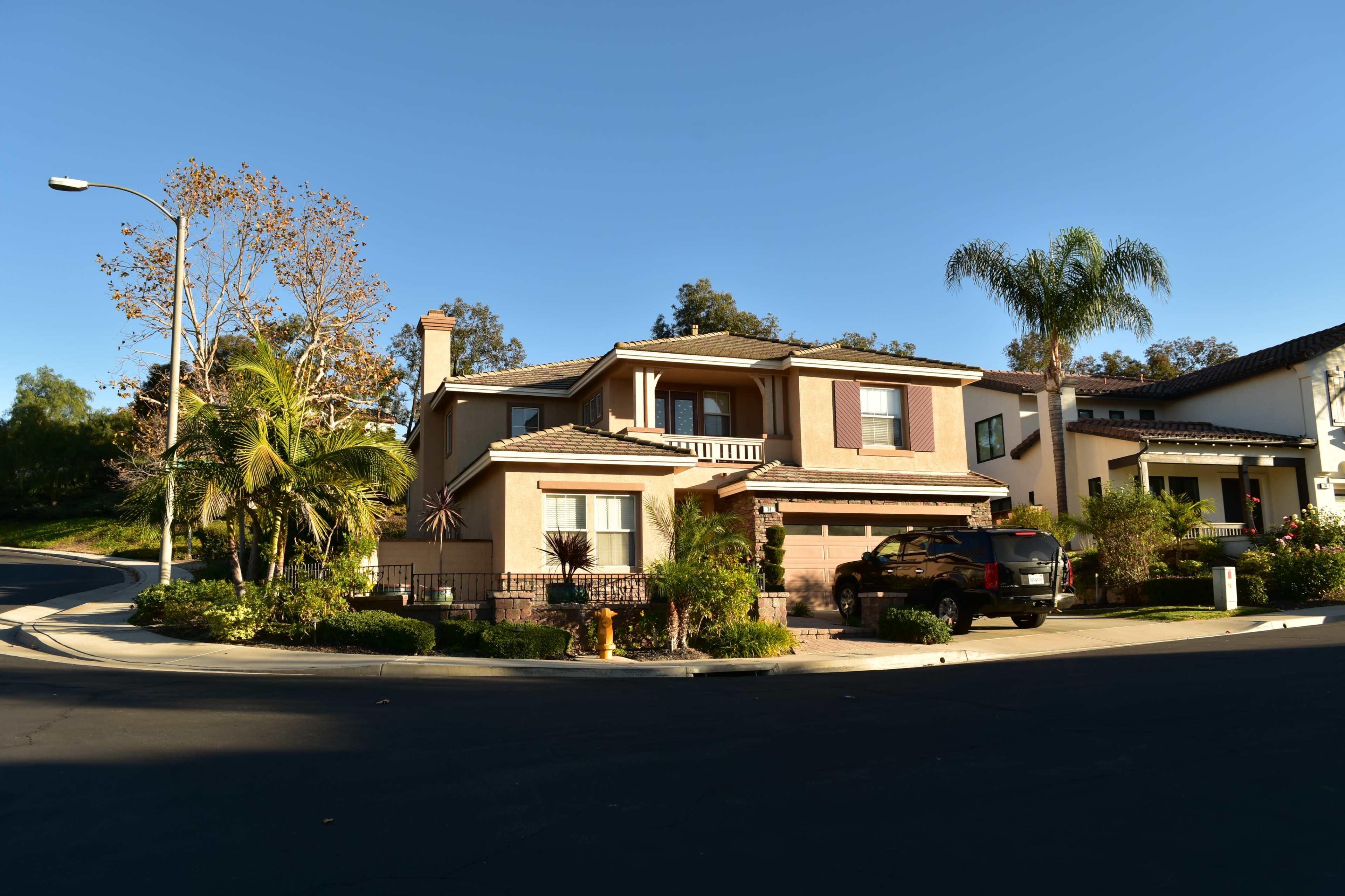 A two-story house with a tiled roof is situated on a corner lot, surrounded by greenery and a well-maintained garden.
