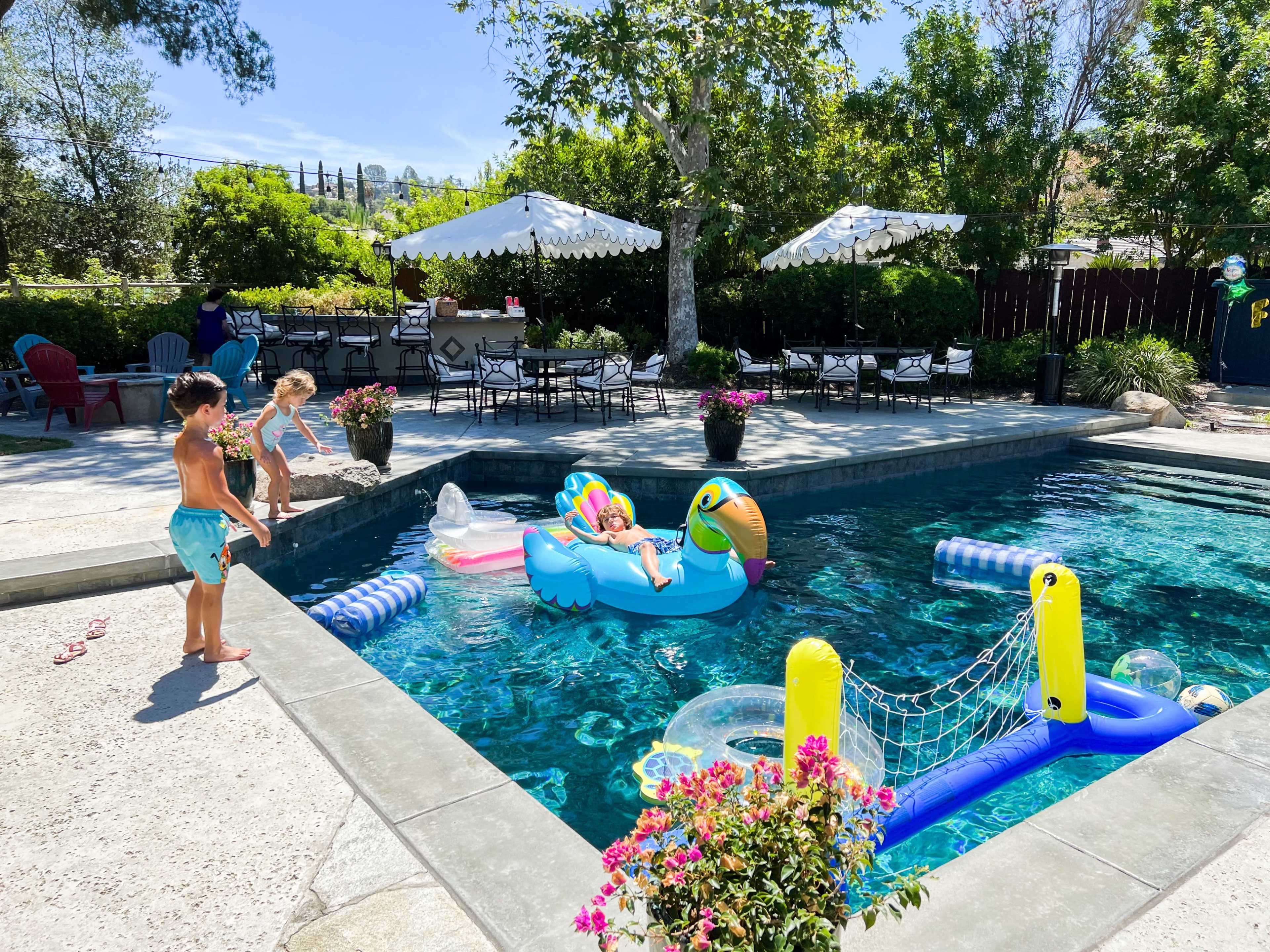 A child stands on the edge of a pool while another child plays with inflatable toys in the water, surrounded by chairs and tables in a sunny backyard.