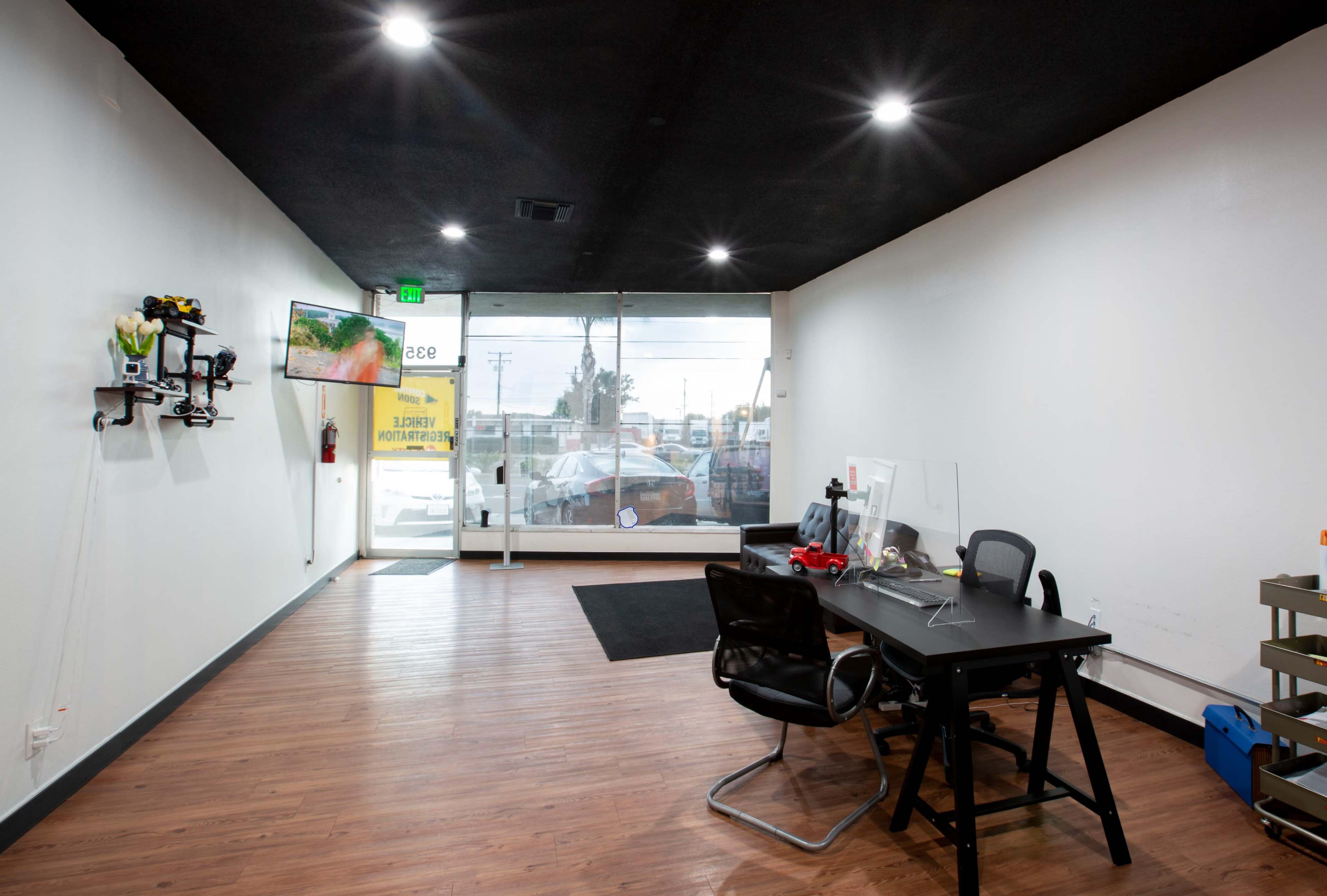 An empty commercial space features a black ceiling, wood flooring, a desk with chairs, and a large window at the entrance.