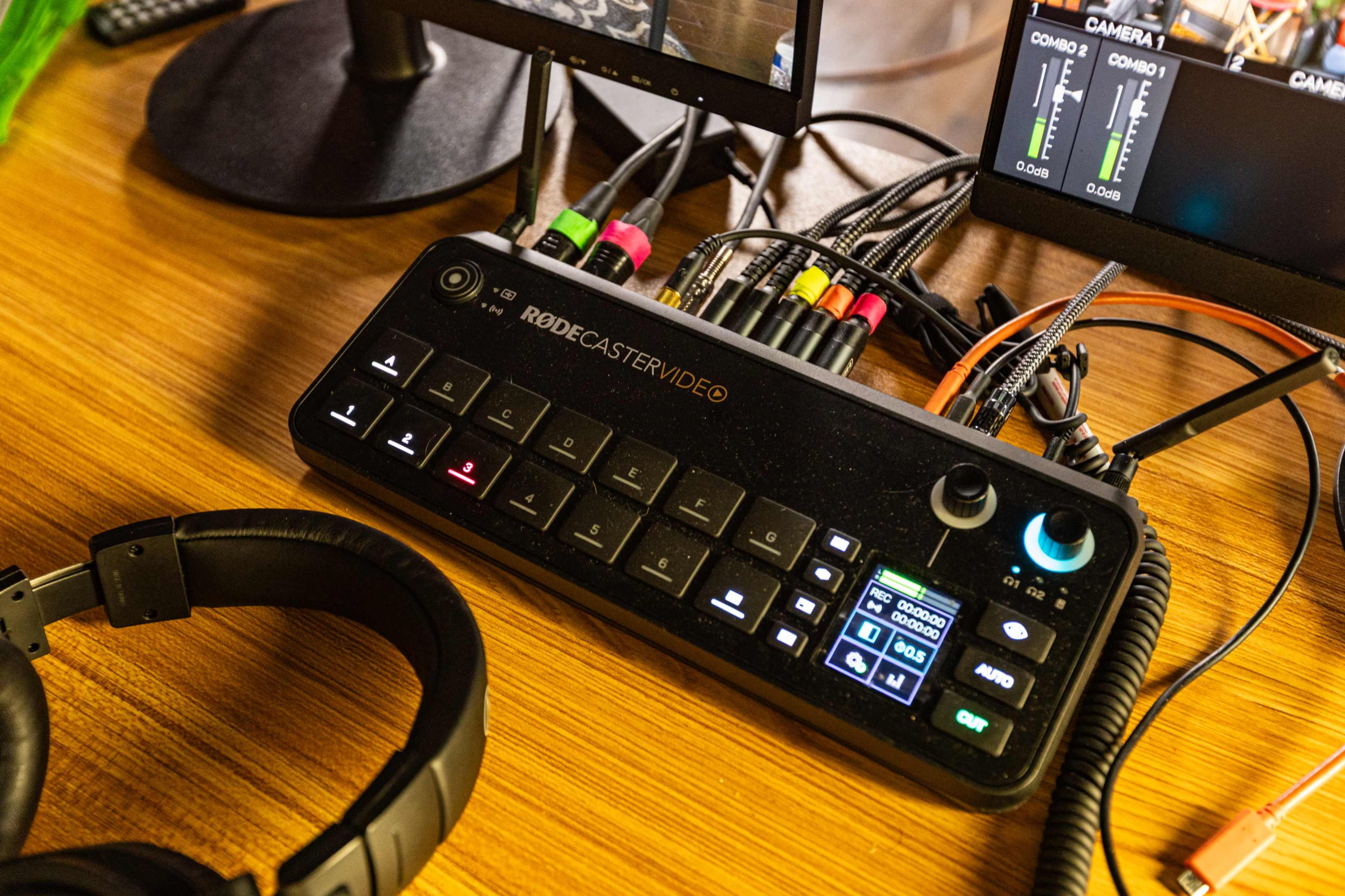The image shows a Rodecaster Pro audio mixer positioned on a wooden surface, with a pair of headphones nearby and several connected cables leading to two computer monitors displaying audio levels.