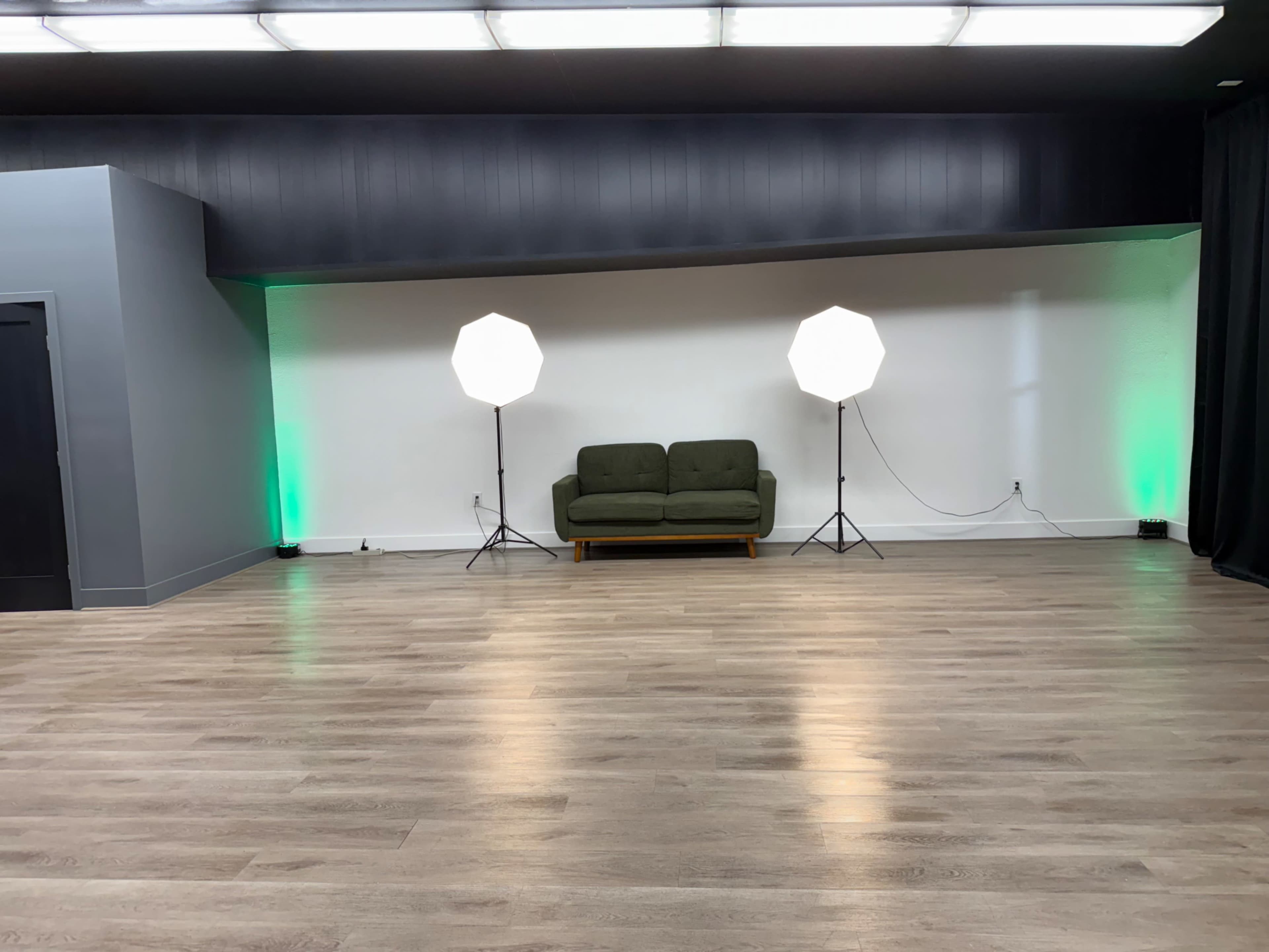 The image shows a spacious room with light wood flooring, featuring a green sofa centered between two light stands with white umbrellas and green accent lighting on the sides.