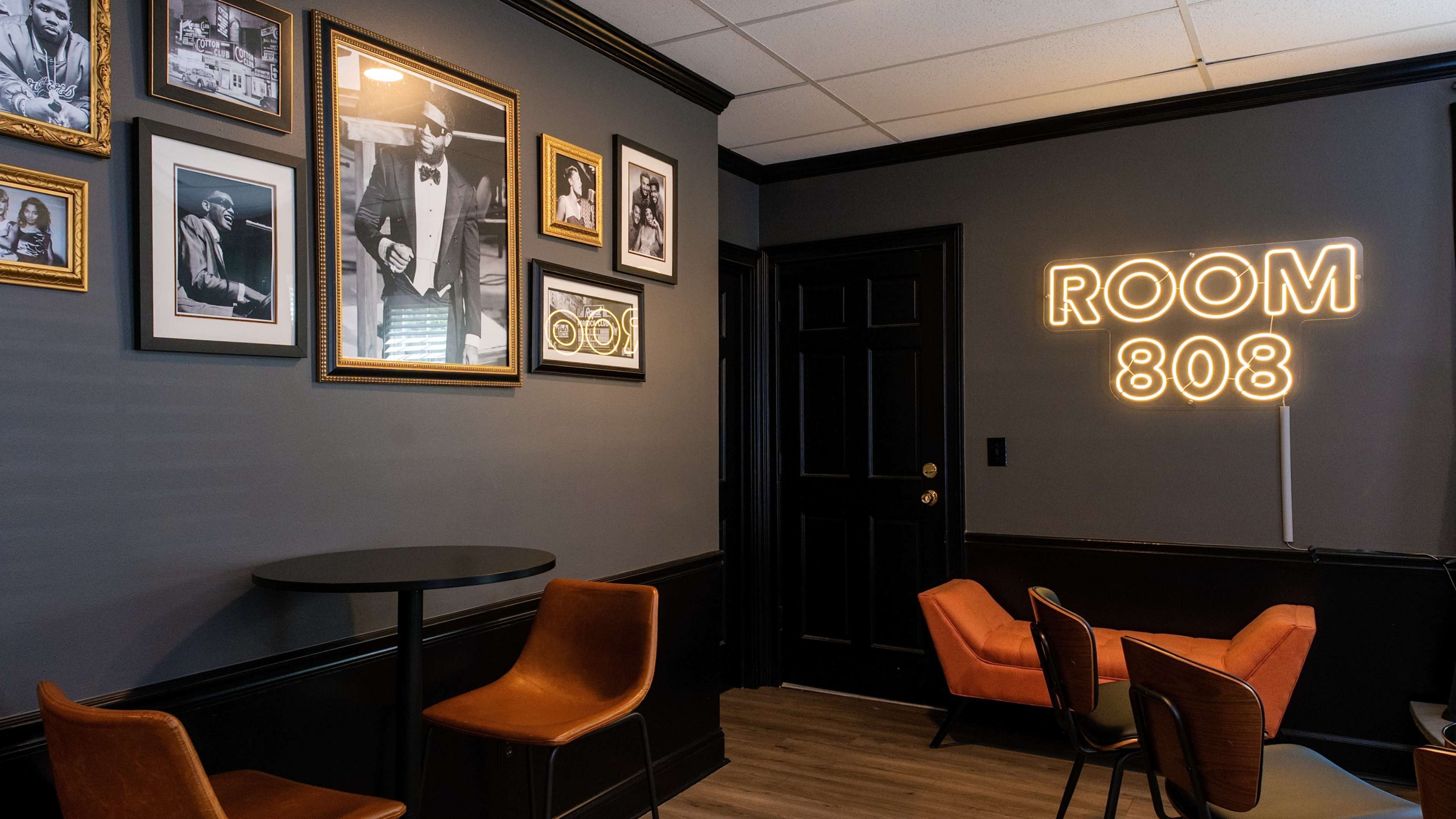 The image shows a stylish room with framed photographs on the walls, a black table, and orange chairs, featuring a neon sign that reads "ROOM 808."