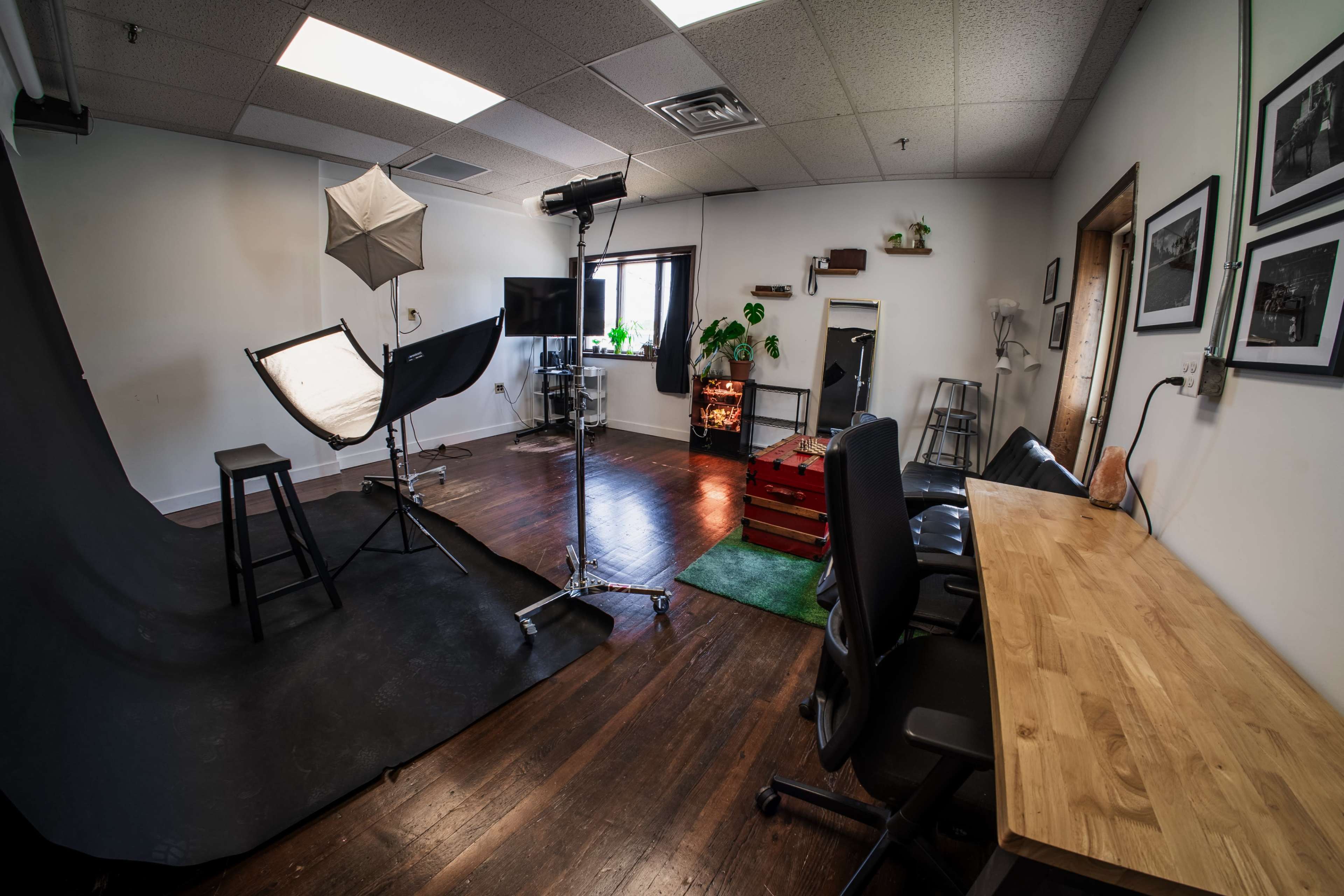 The image shows a photography studio with a backdrop, lighting equipment, a small seating area, and a desk arranged against a wall with decorative plants.