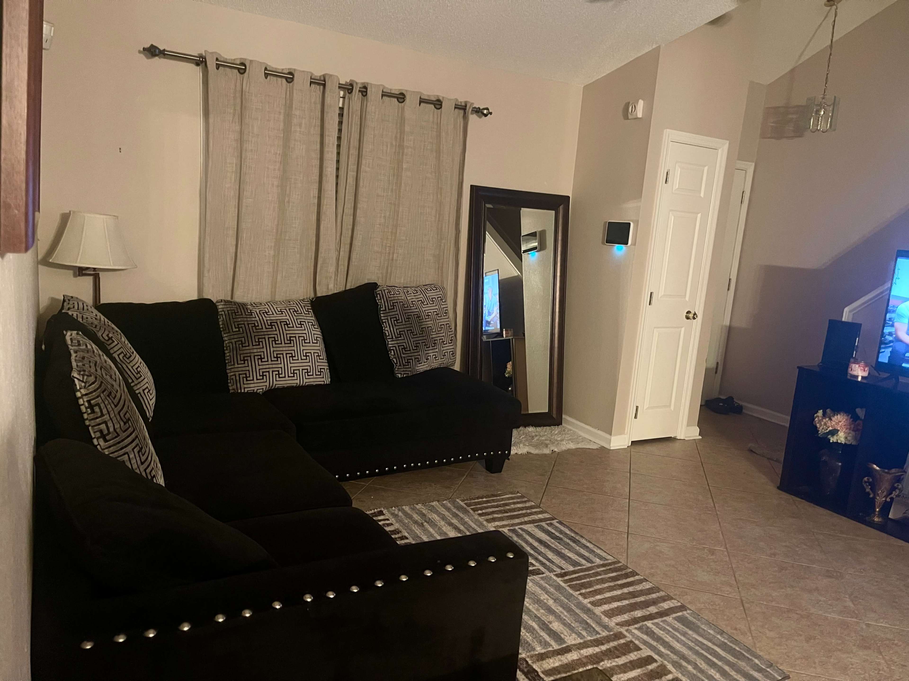 A living room with a black sectional sofa, a large mirror, and a television on a stand against a beige wall.