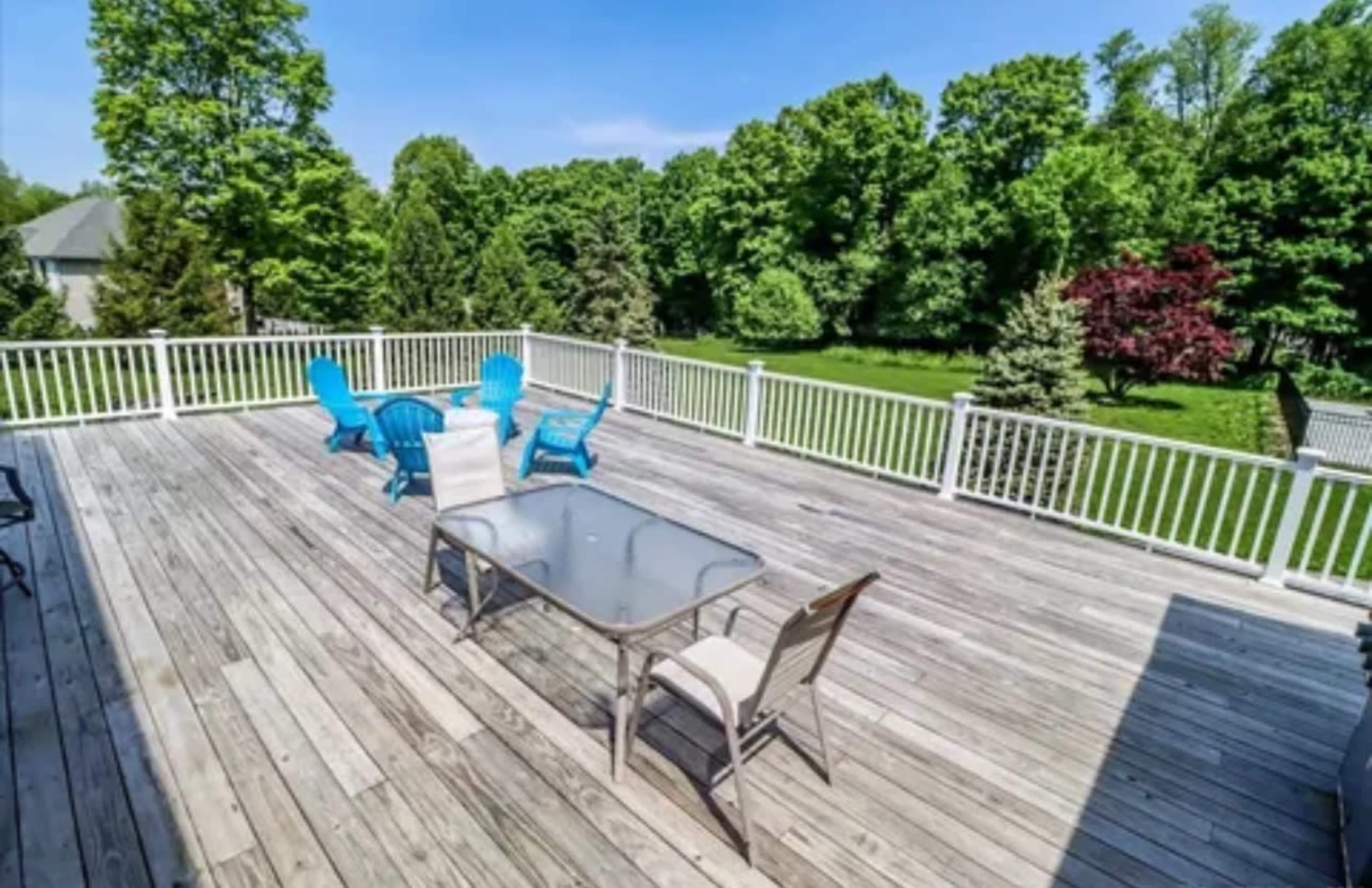 A spacious wooden deck features a glass-top table and two blue lounge chairs, surrounded by lush greenery.