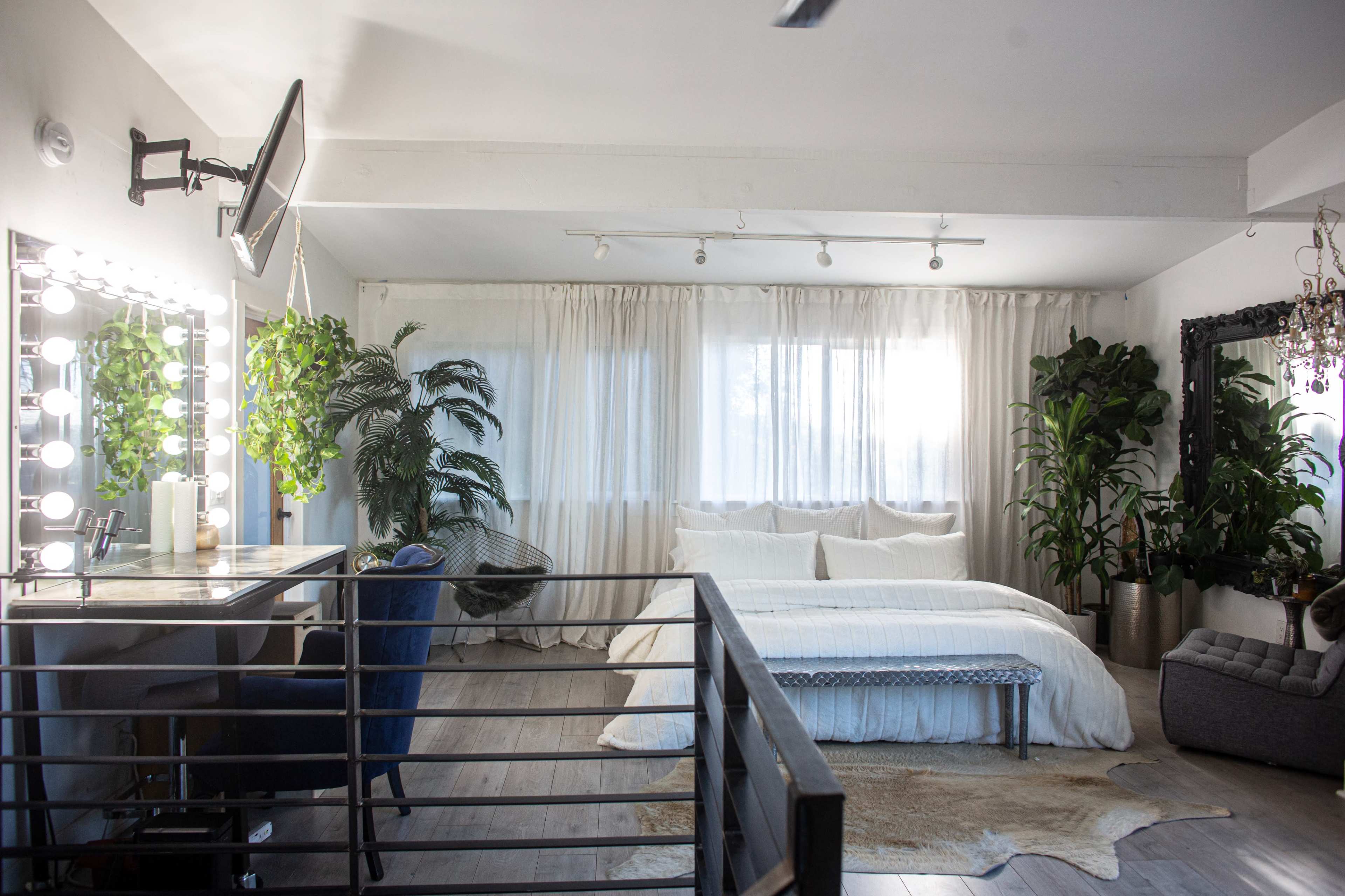 A spacious bedroom features a large bed, a vanity with lights, and several indoor plants beside large windows with sheer curtains.