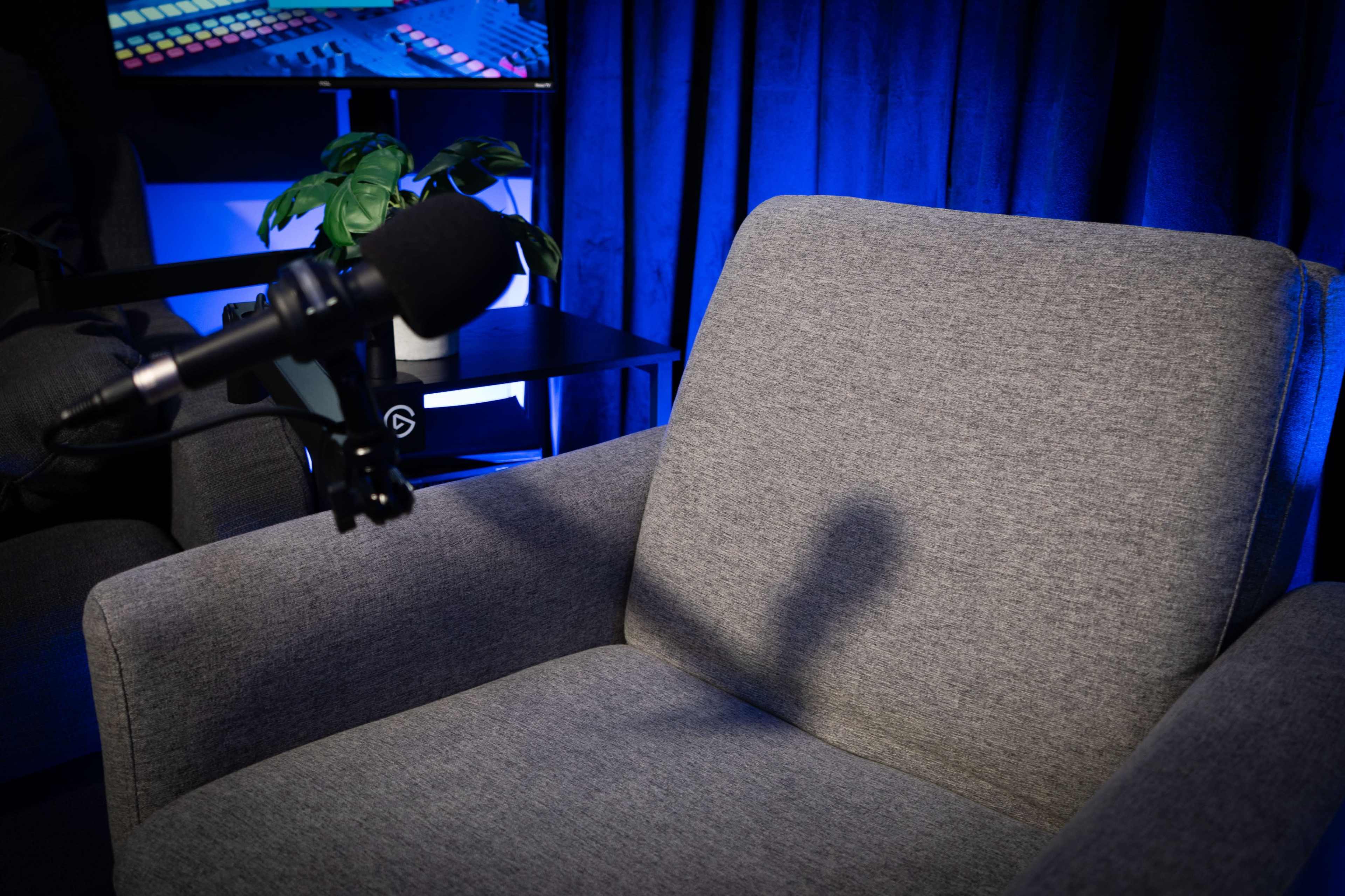 The image shows a gray armchair with a microphone positioned in front of it, and a plant in a pot nearby against a backdrop of blue lighting.