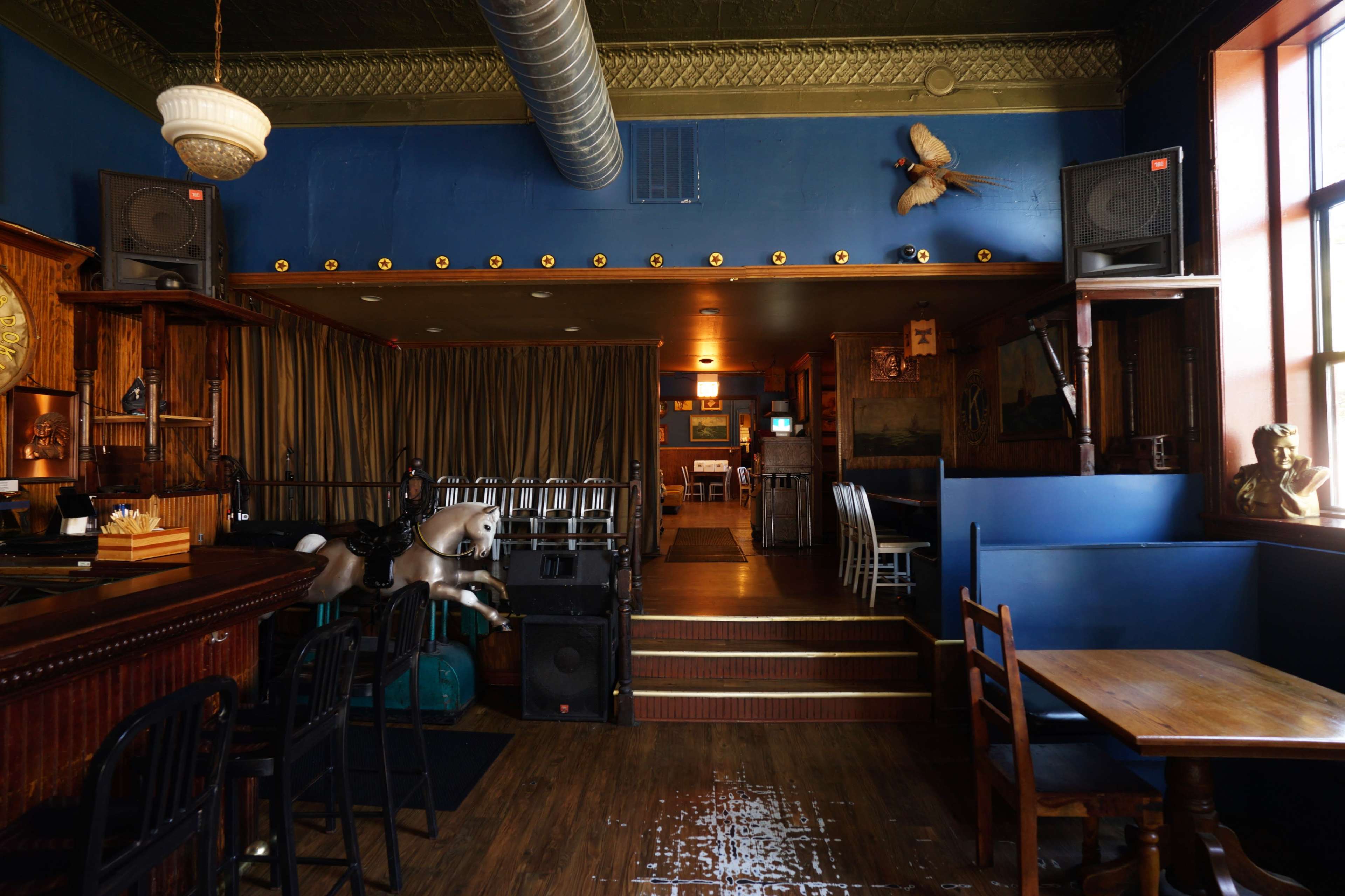The interior of a bar with a wooden counter, a horse statue, seating areas, and audio equipment arranged against blue walls.