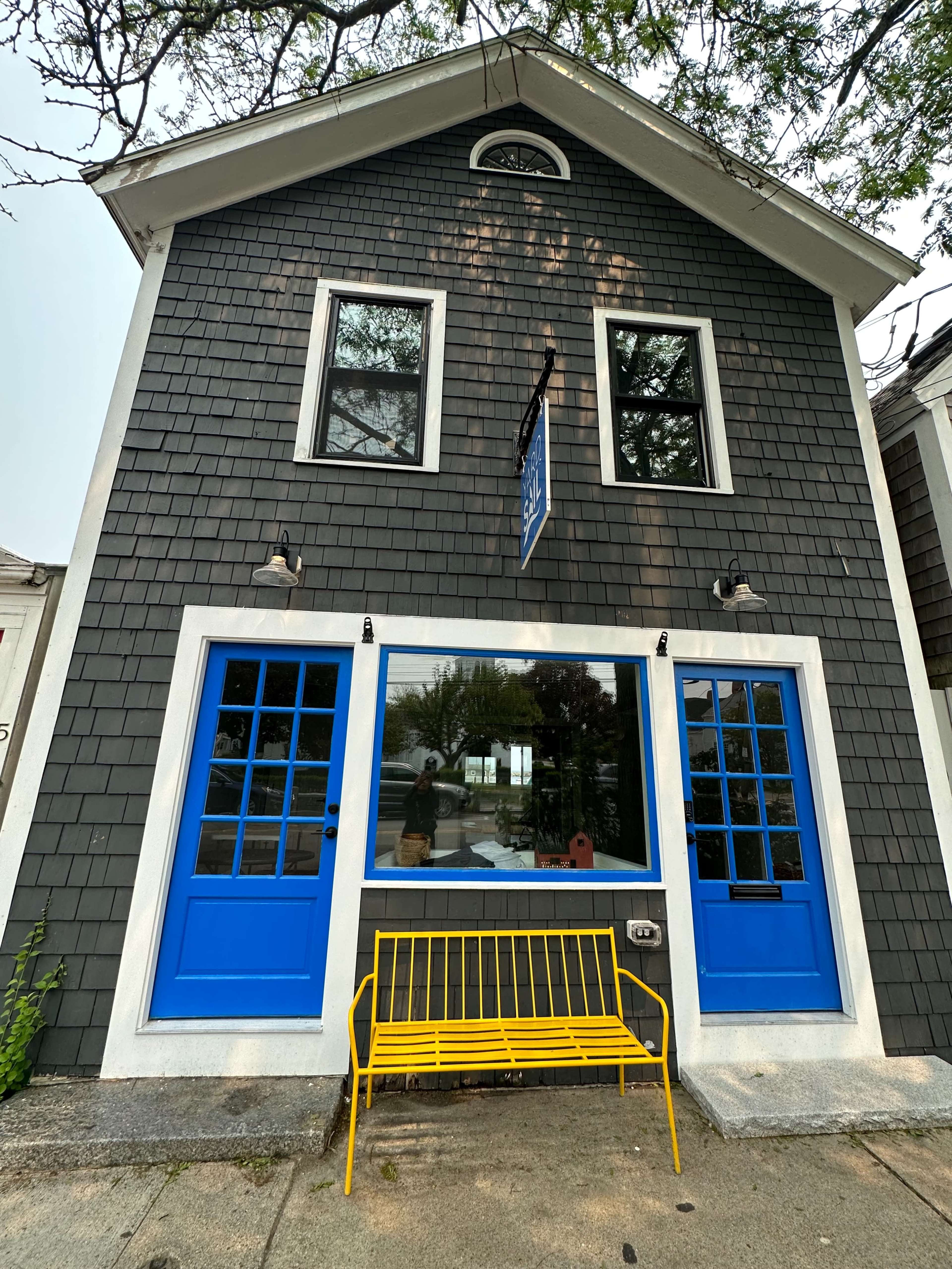 A two-story gray shingle building with bright blue double doors, a large central window, and a yellow bench in front.