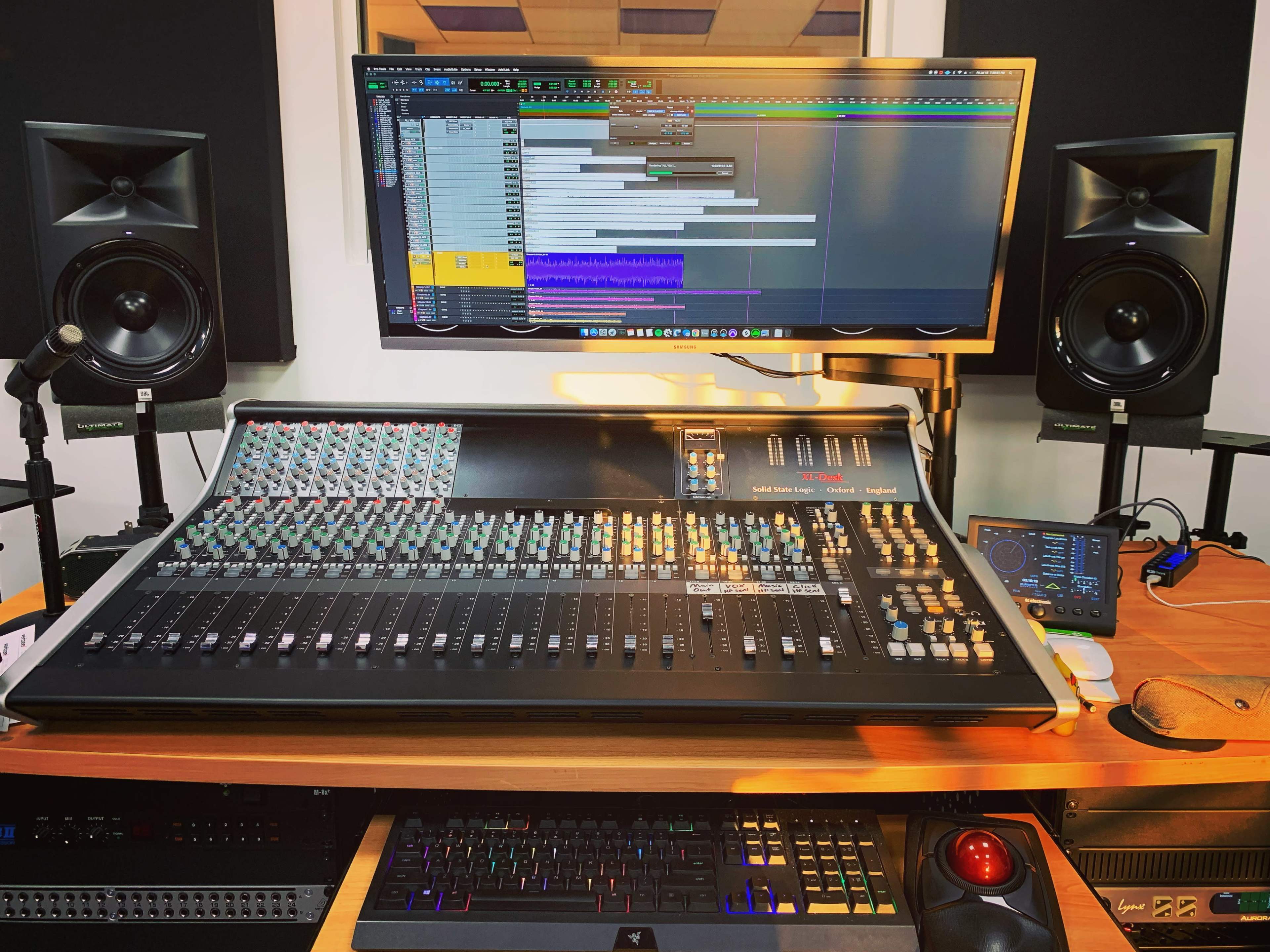 A digital audio workstation setup features a mixing console, computer monitors displaying audio tracks, and studio monitors on either side.