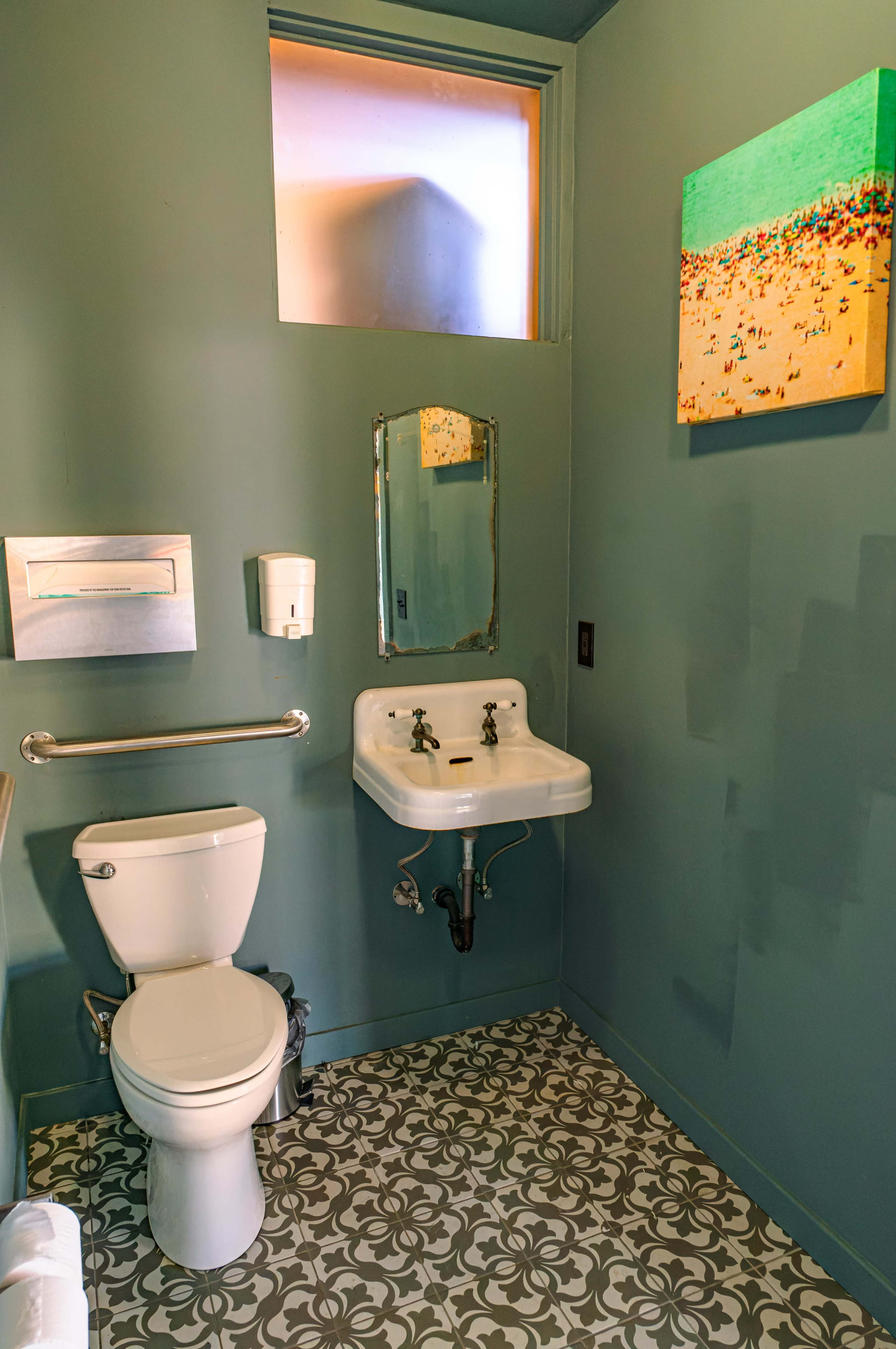 A small bathroom featuring a toilet, a wall-mounted sink, a mirror, and a colorful painting on the wall, with patterned tiles on the floor.