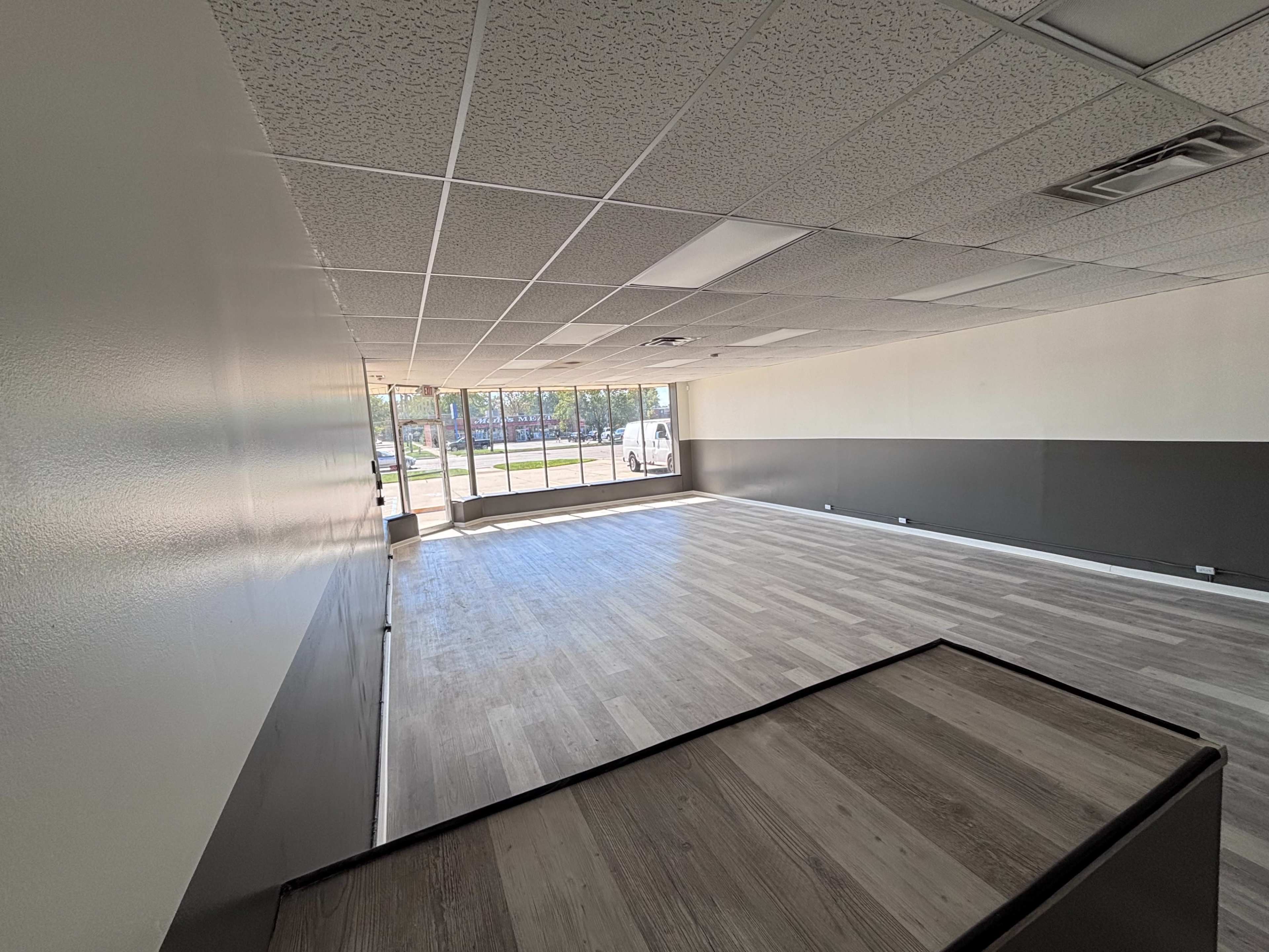 The image shows an empty commercial space with light-colored walls, a wooden floor, and large windows letting in natural light.