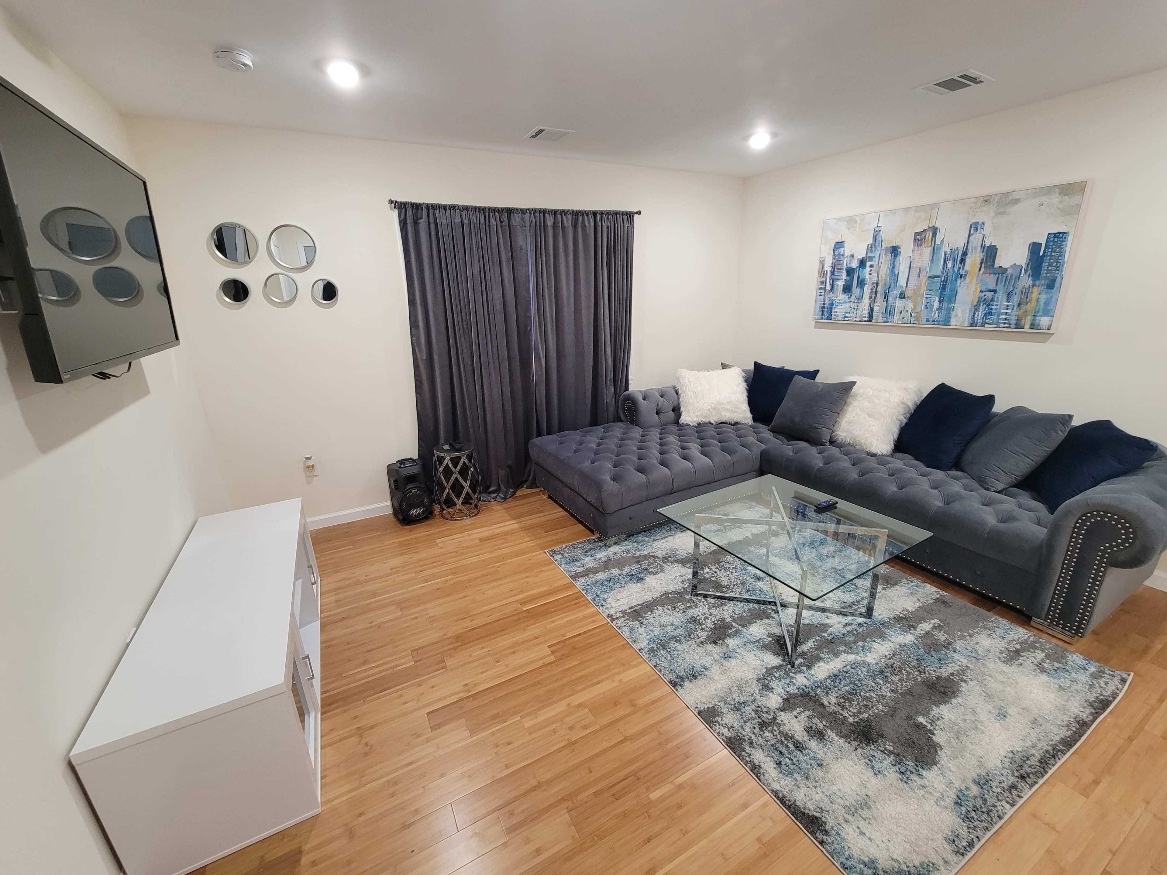 The room features a sectional sofa, a glass coffee table, a wall-mounted TV, a curtain, and decorative elements, all arranged on a hardwood floor.