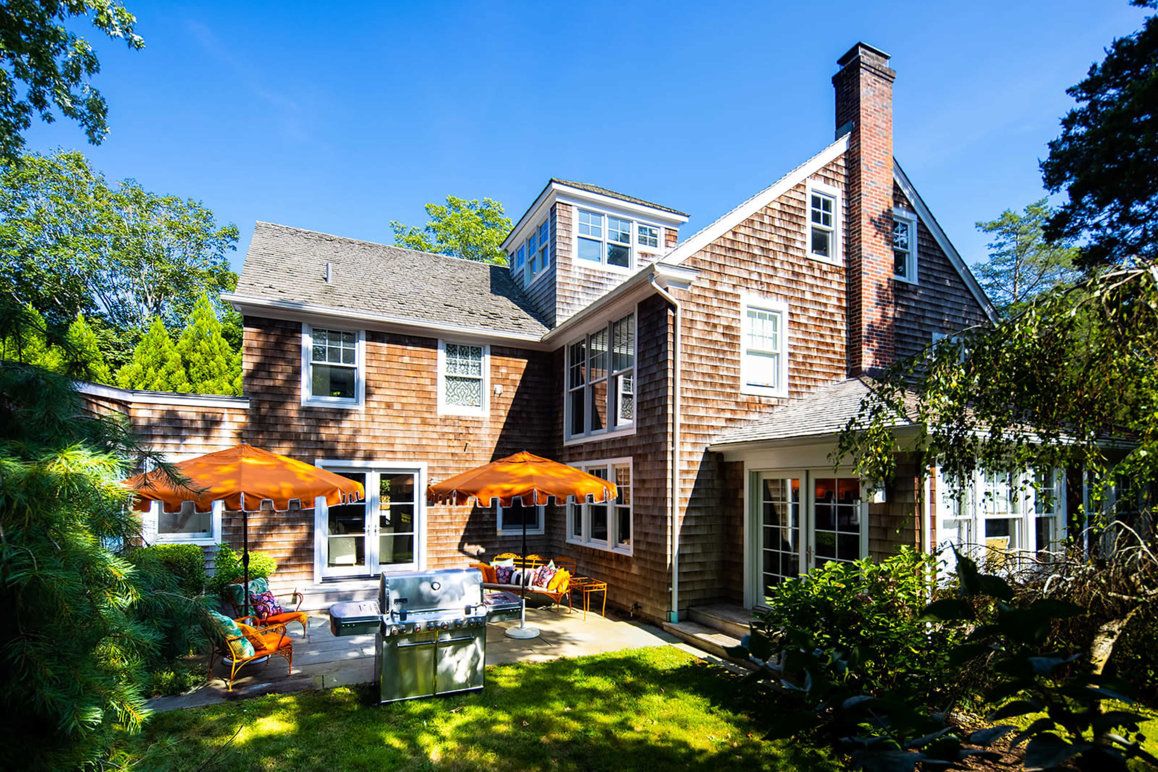 A two-story wooden house with a patio, grill, and orange umbrellas sits in a green yard under clear blue skies.