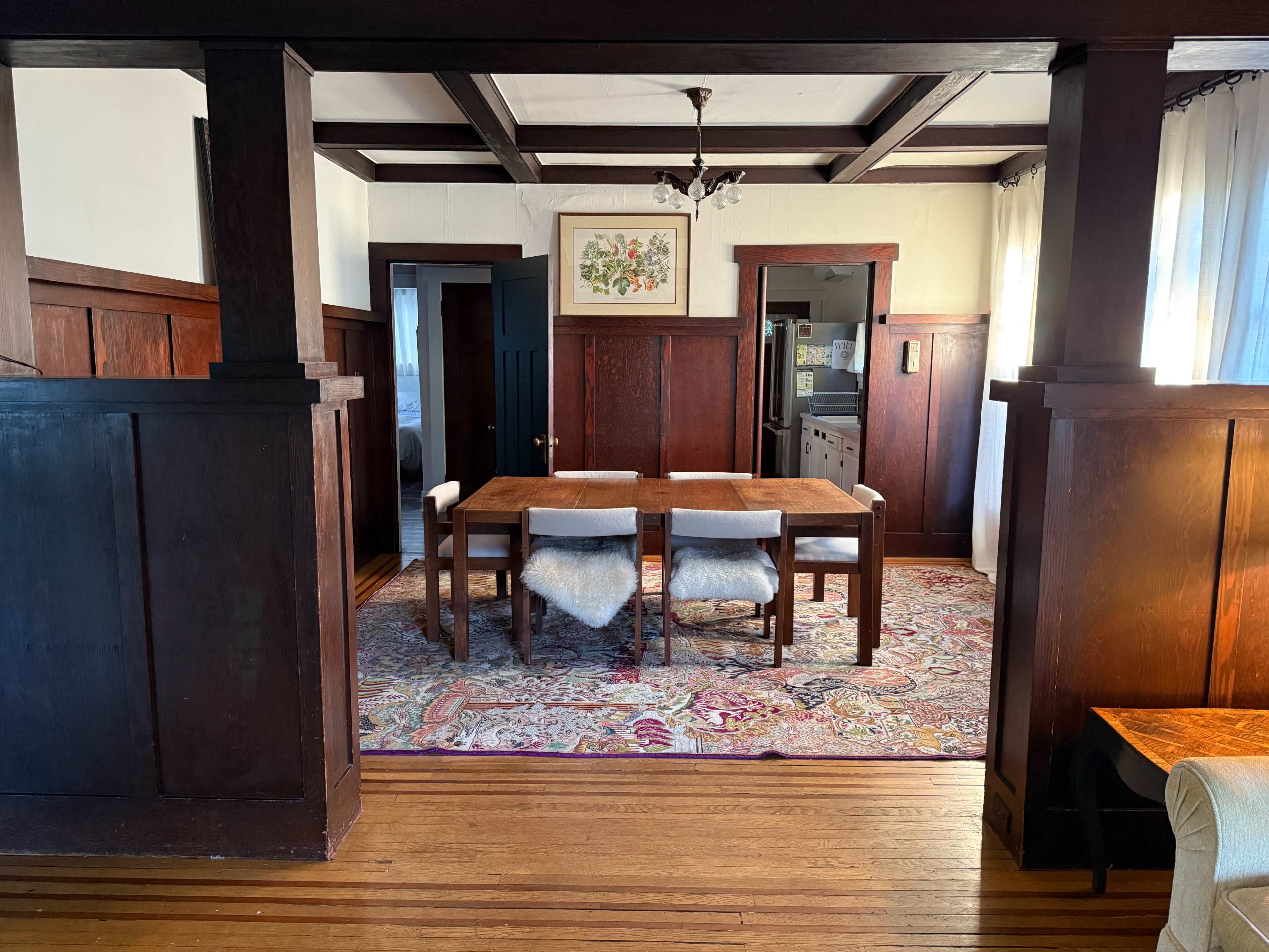 The image shows a dining room featuring a wooden table with chairs, a patterned rug, and dark wood paneling, leading to a kitchen in the background.