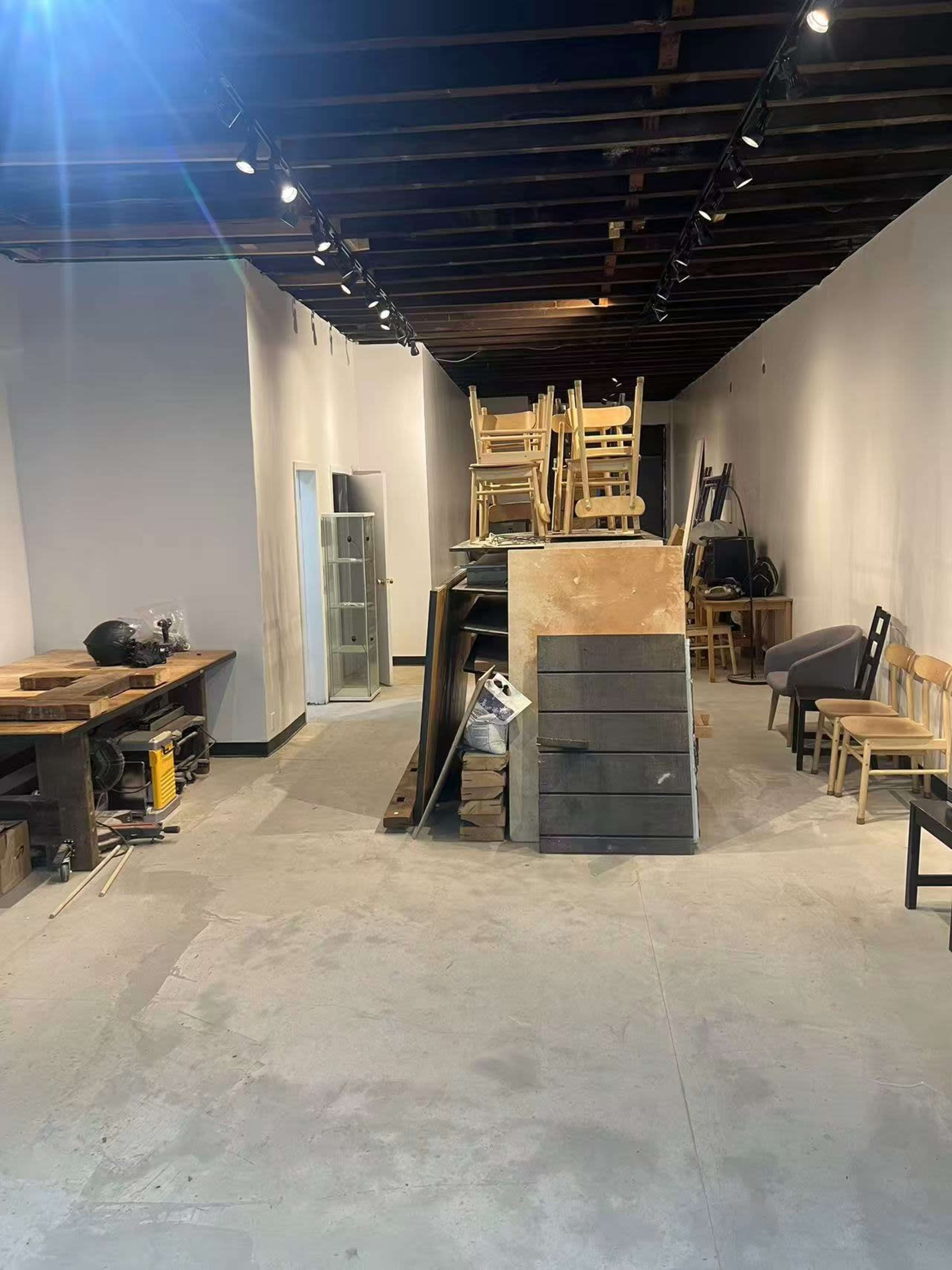 The image shows an empty room with a stack of wooden chairs and various construction materials in the center, along with some furniture and tools along the walls.