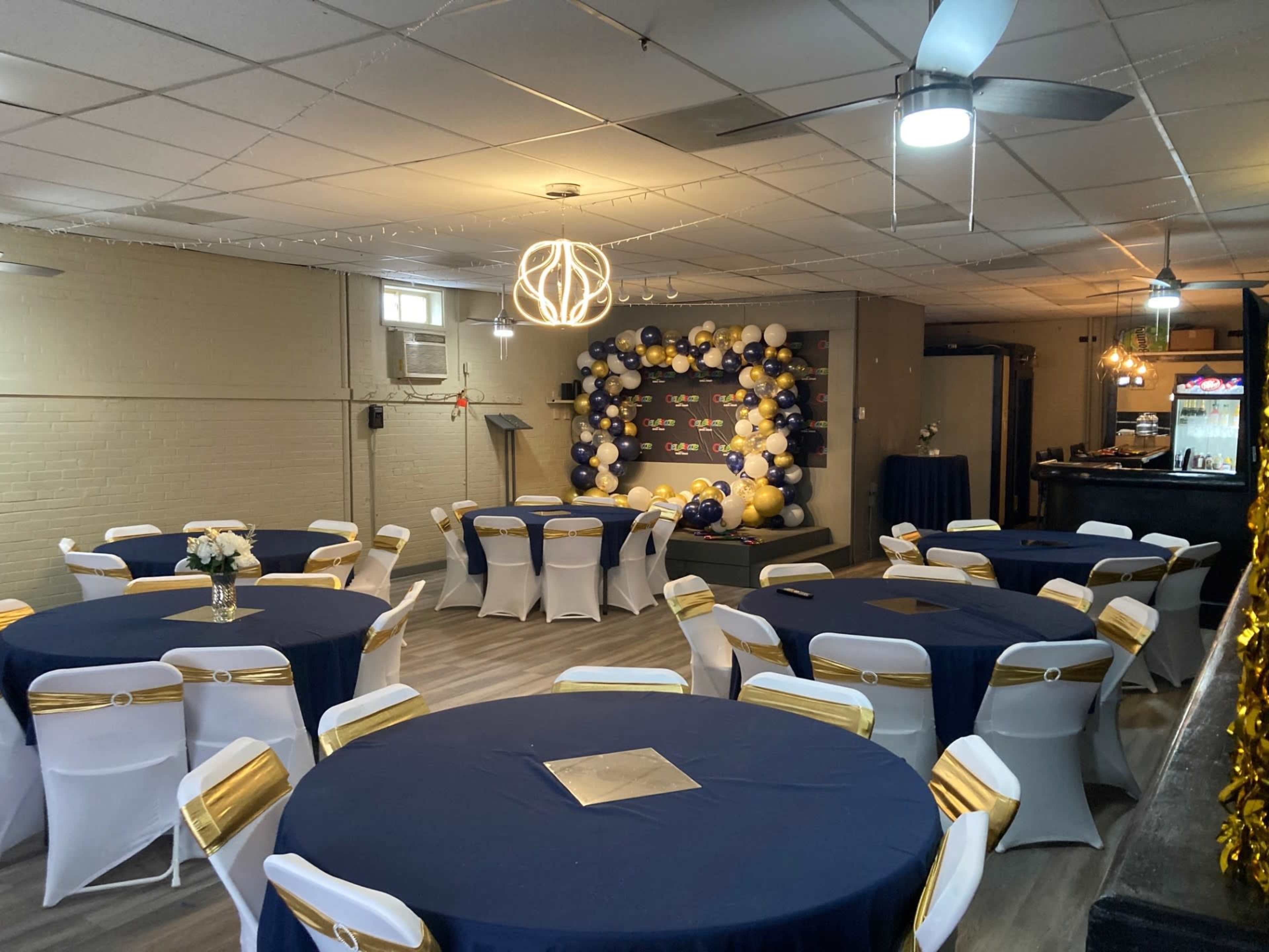The room is set up for an event with round tables covered in blue tablecloths and white chairs adorned with gold sashes, featuring a backdrop of balloons in various colors.