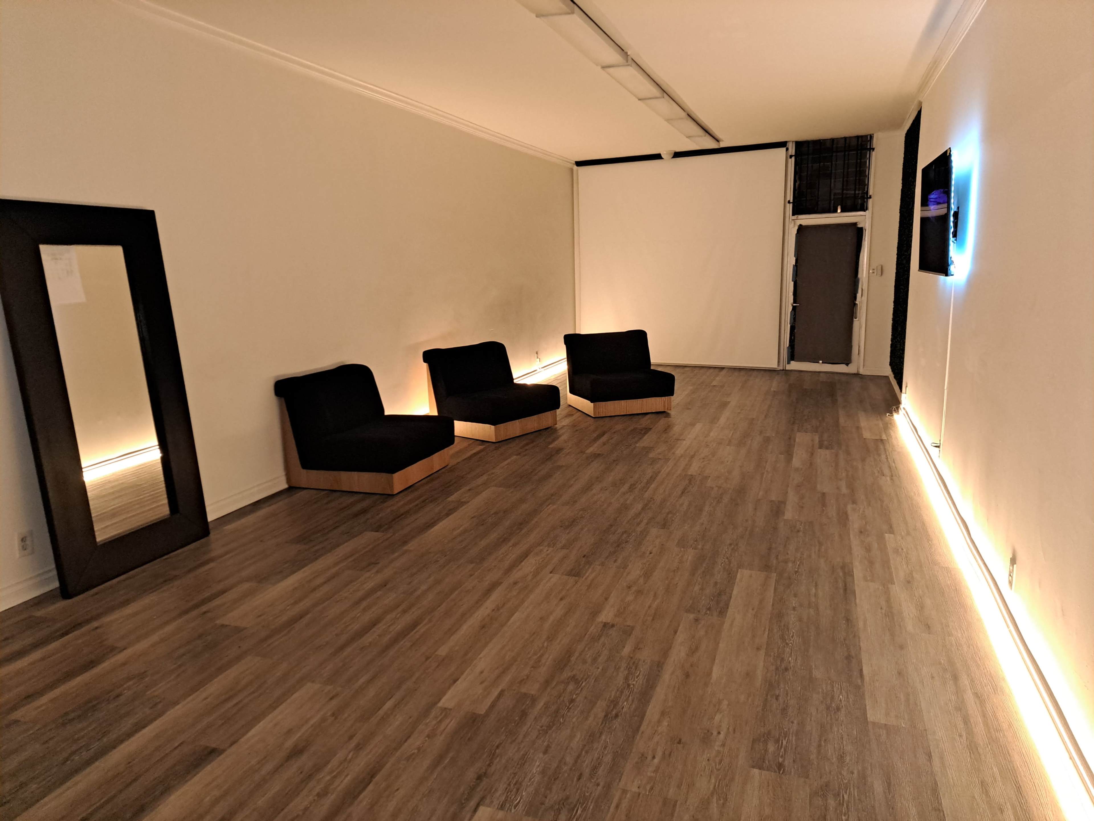 A minimalist room with three black chairs positioned on a wooden floor, a large mirror on the left wall, and sparse lighting.