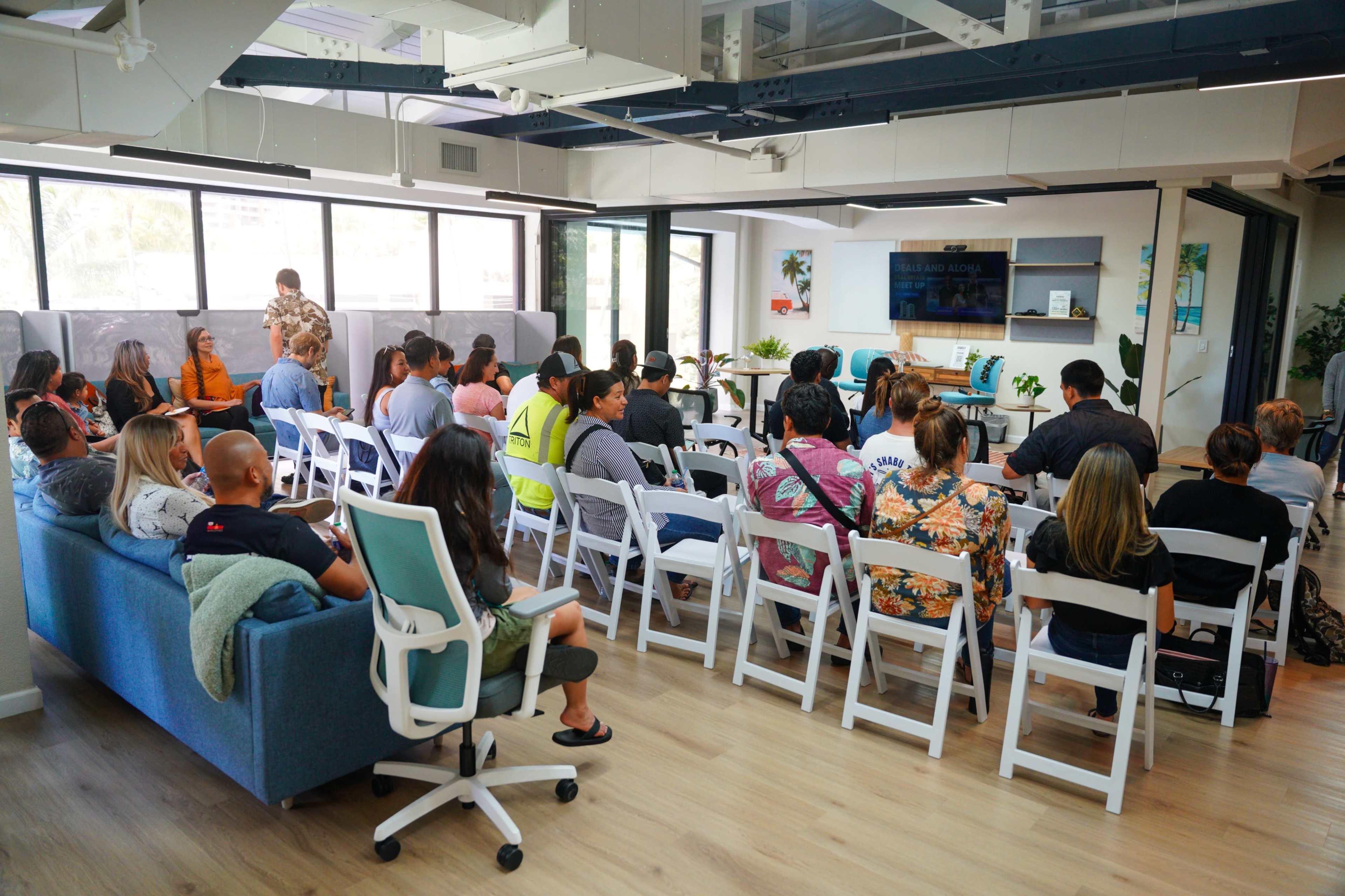 A group of people sits in rows of chairs facing a presenter in a modern office space with large windows and colorful decor.
