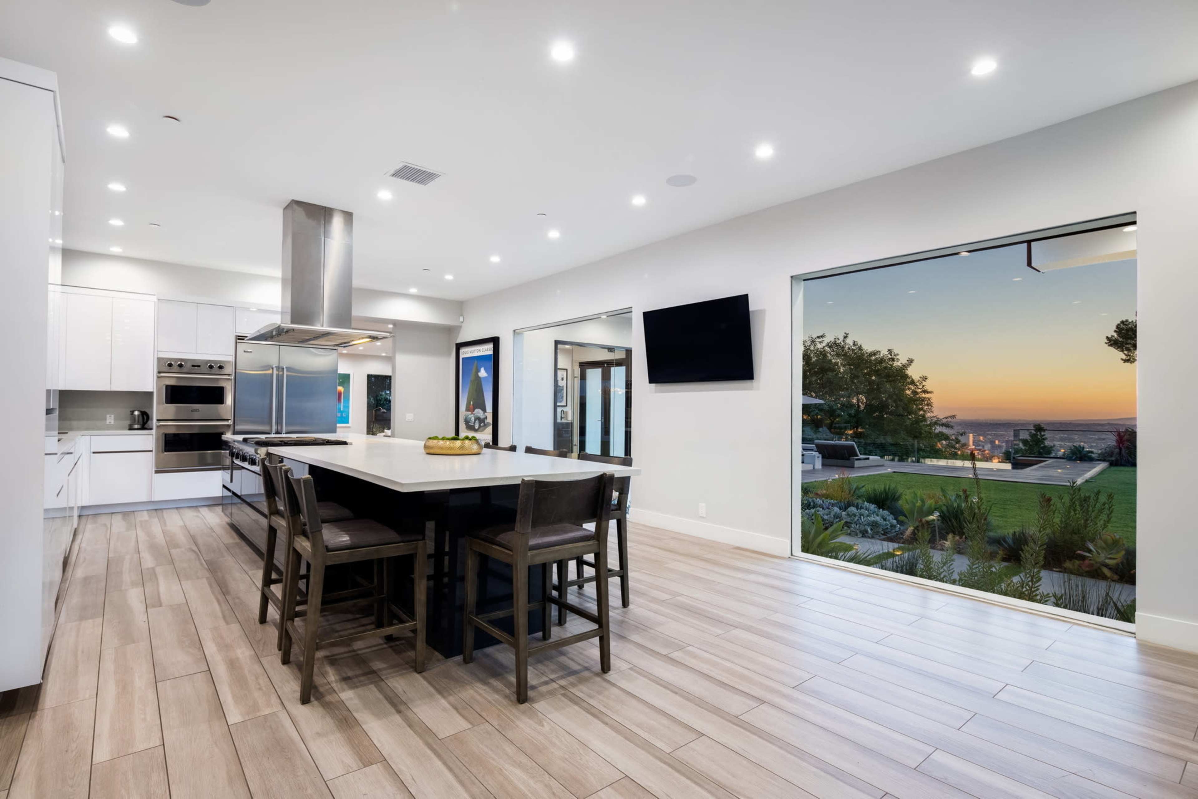 A modern kitchen features sleek white cabinets, a large island, and opens to a patio showcasing a sunset view.
