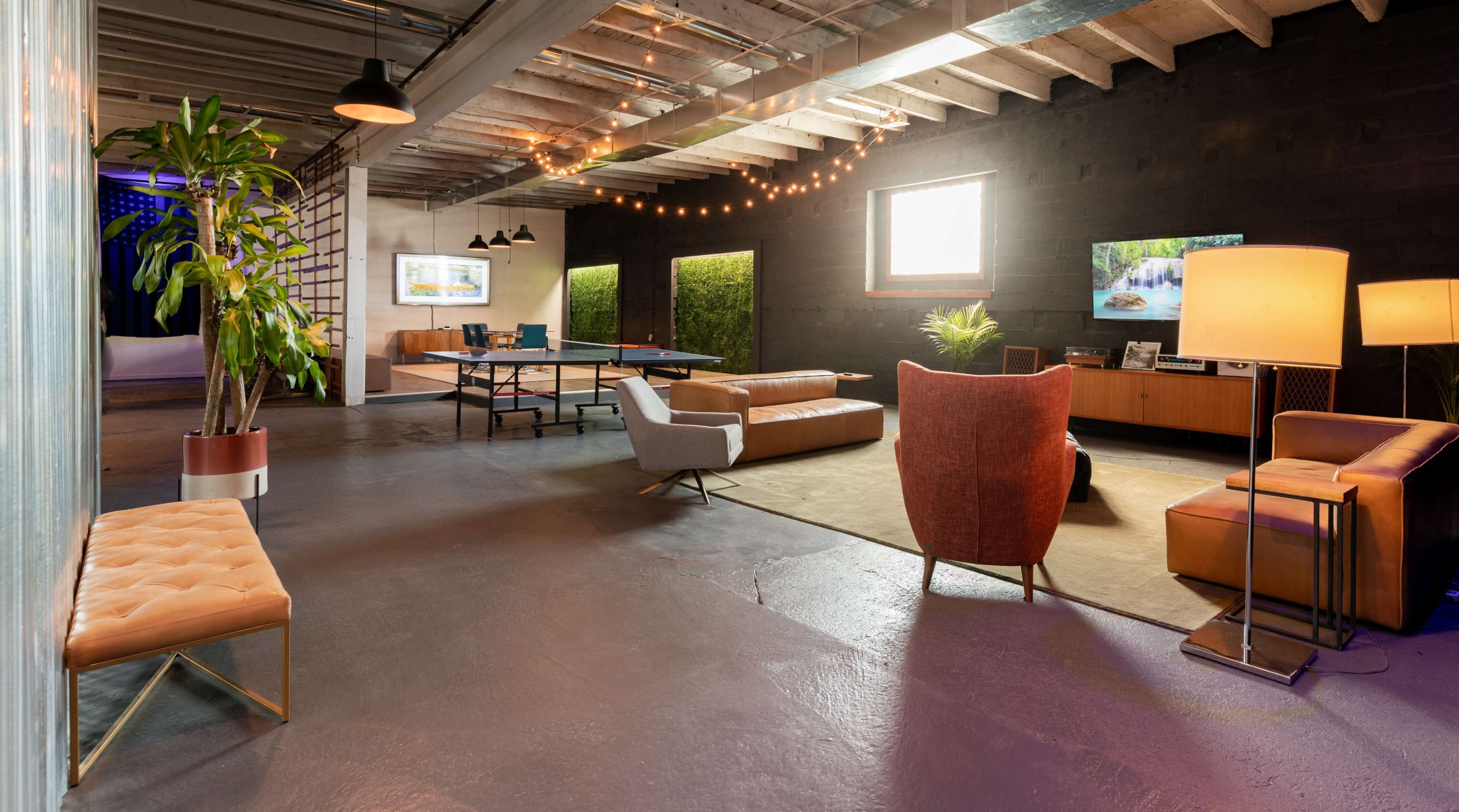 The image shows a modern, industrial-style lounge area featuring a mix of seating, a ping pong table, and decorative plants.