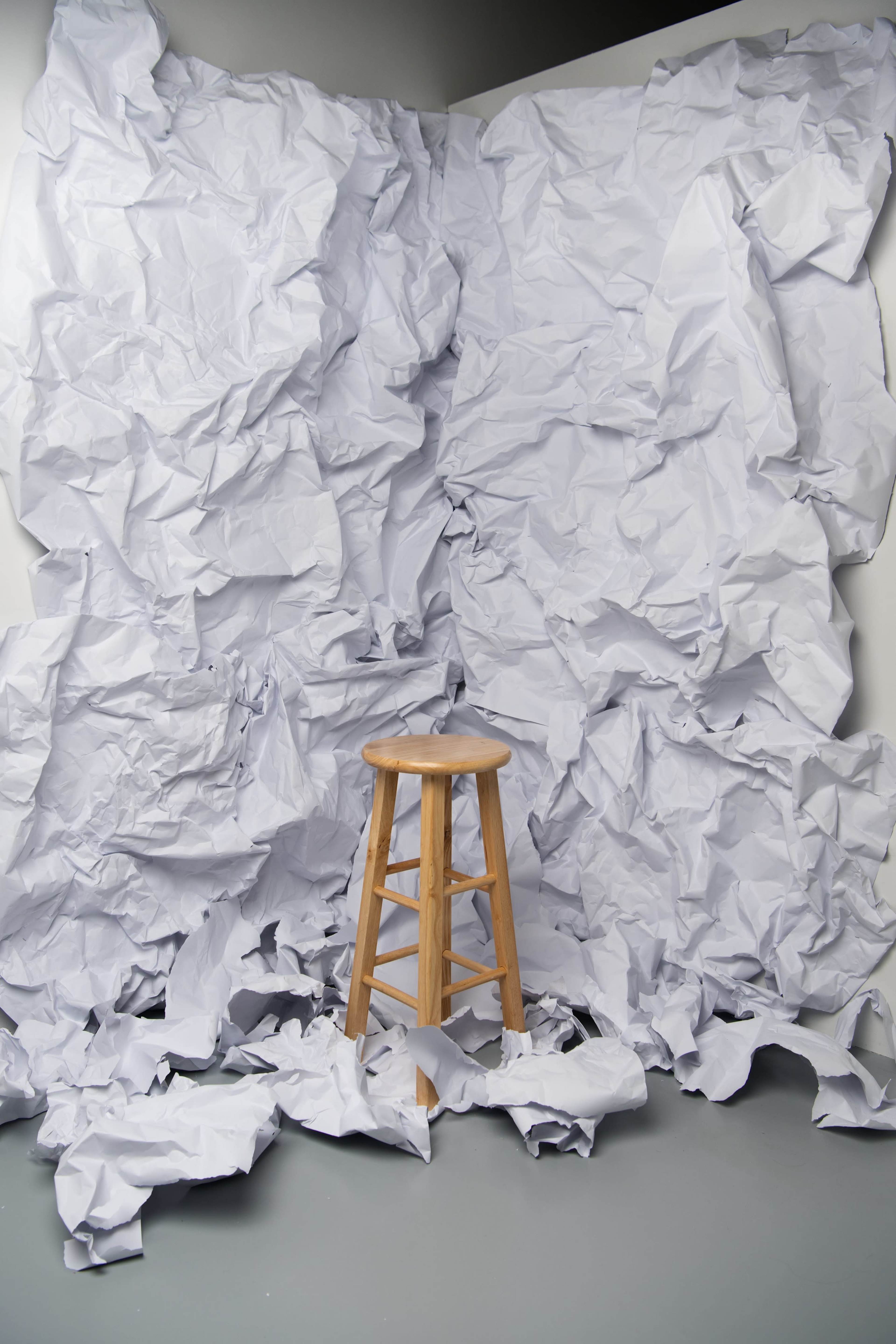 A wooden stool stands in front of a textured backdrop made of crumpled white paper.