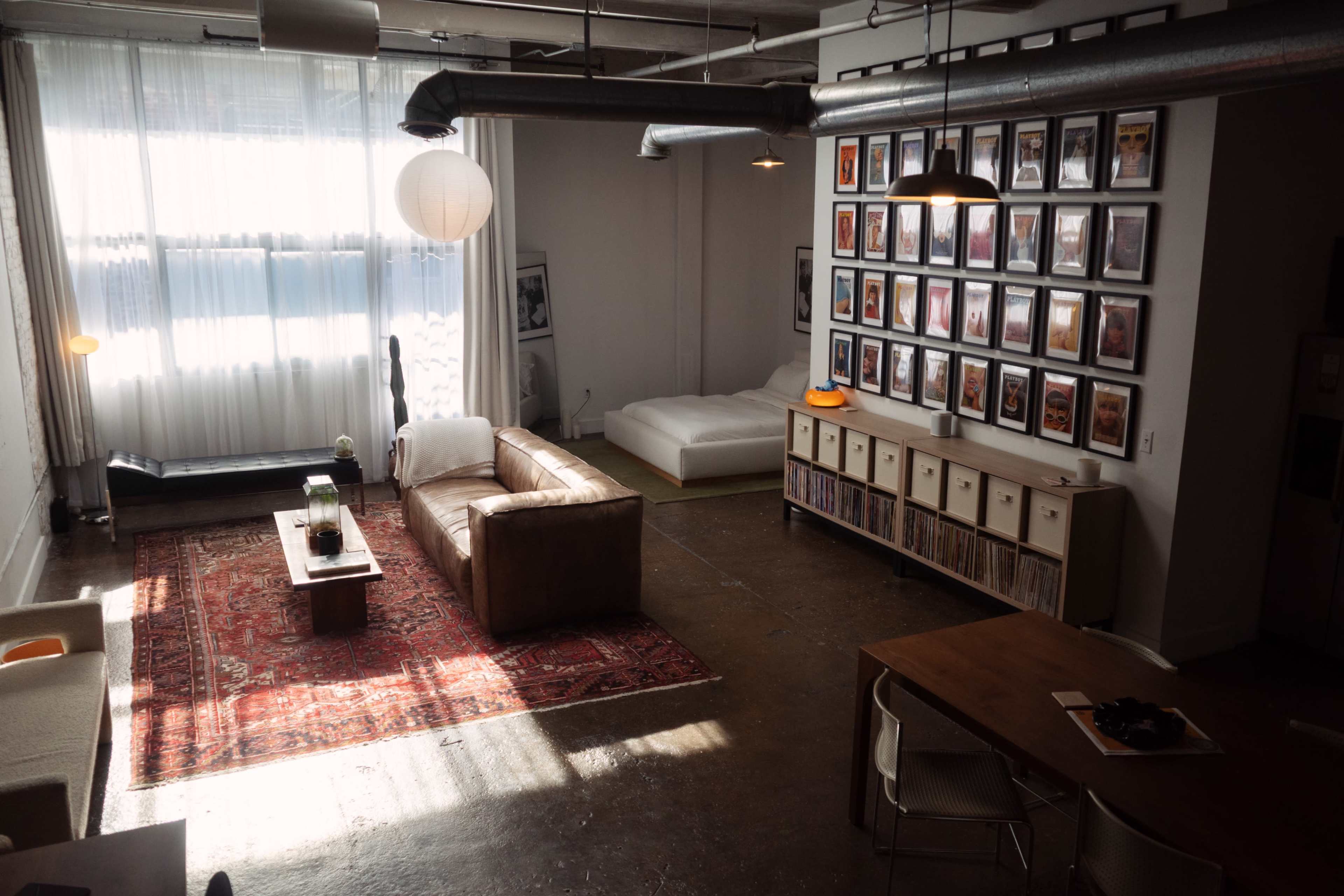The image shows a spacious, minimally furnished living area featuring a large sofa, a wooden coffee table, a dining table, and walls adorned with framed artwork.