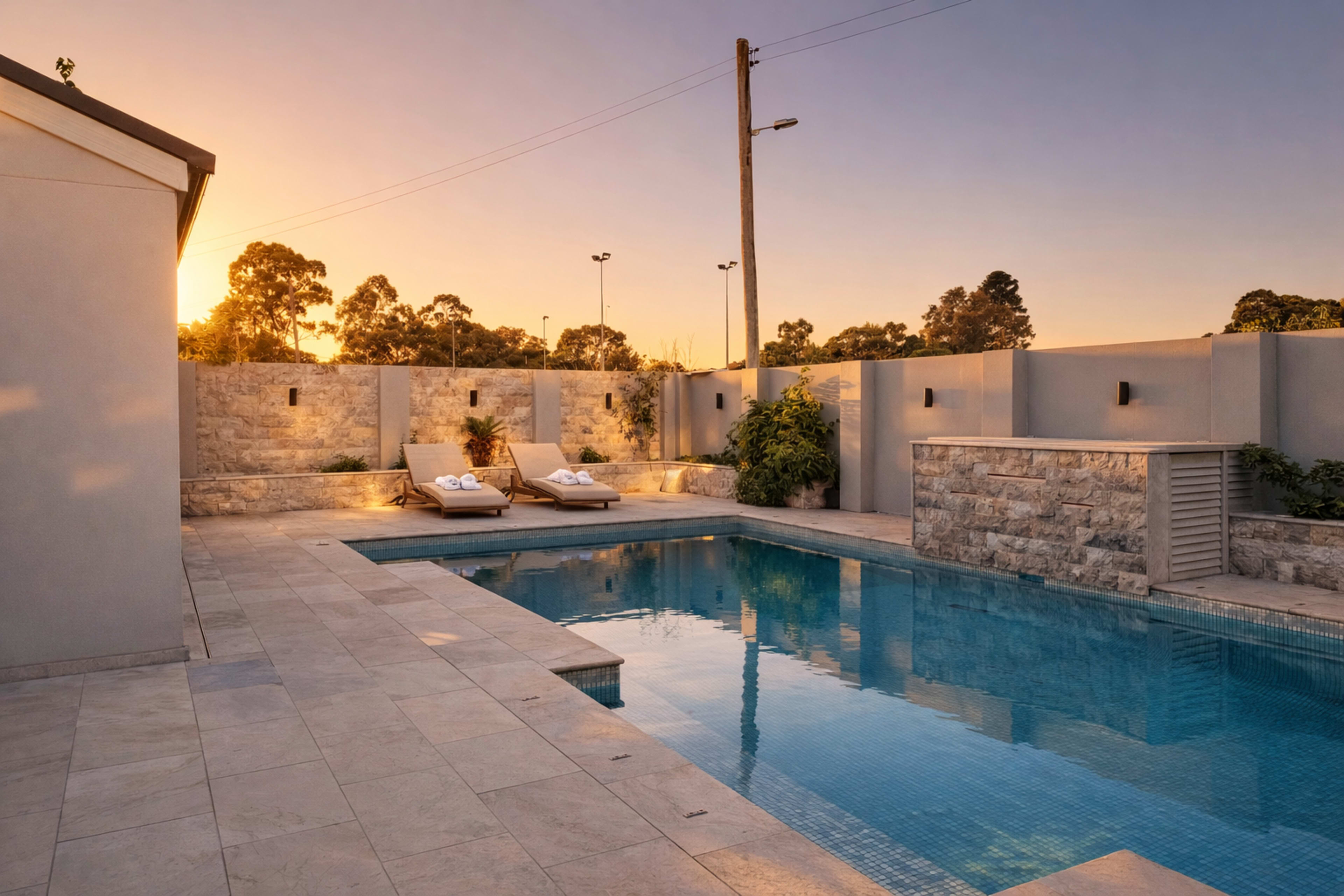 The image shows a modern outdoor pool area featuring two lounge chairs, stone walls, and a sunset in the background.