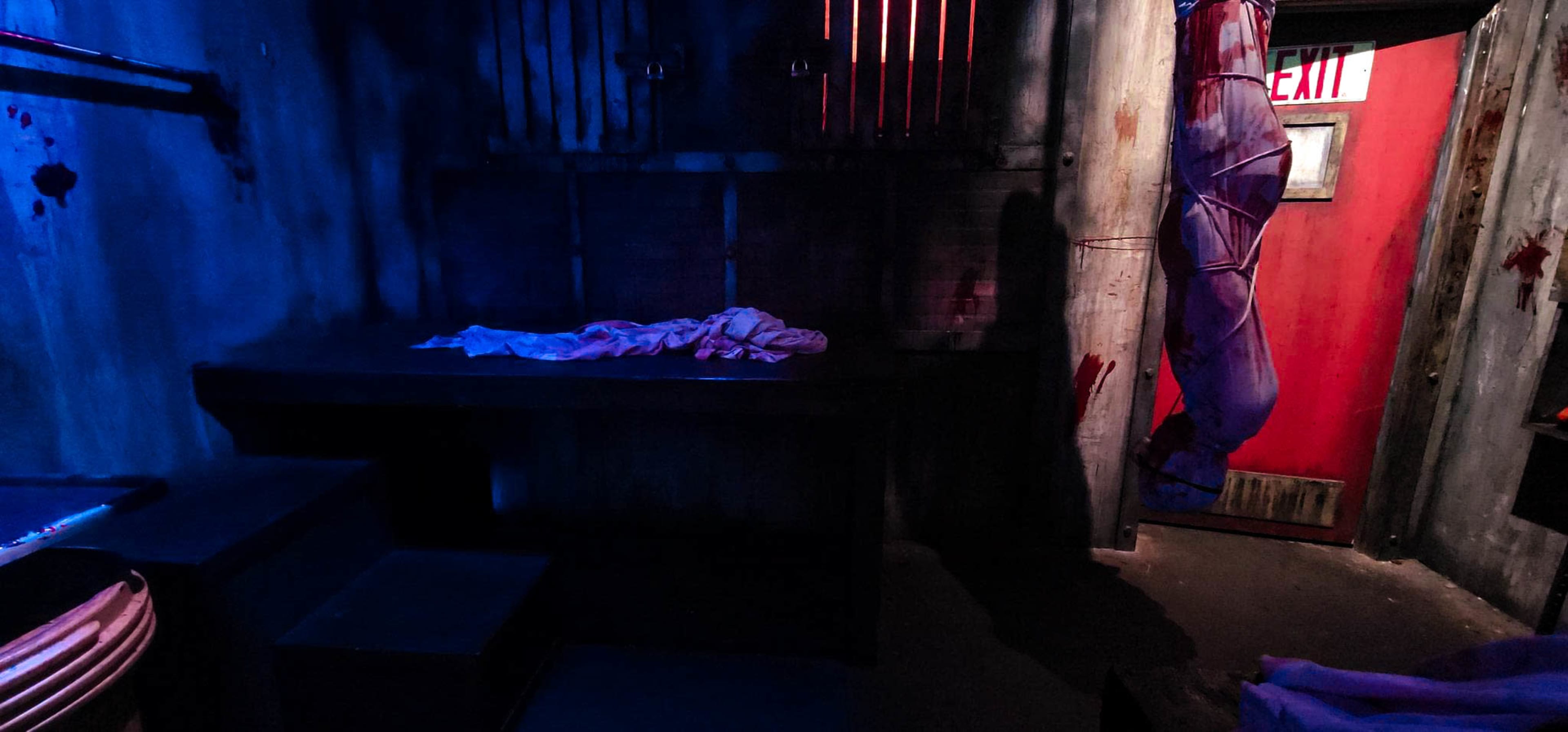 The dimly lit room features a bloodstained wall, a table with a white cloth, and a door marked with an exit sign.