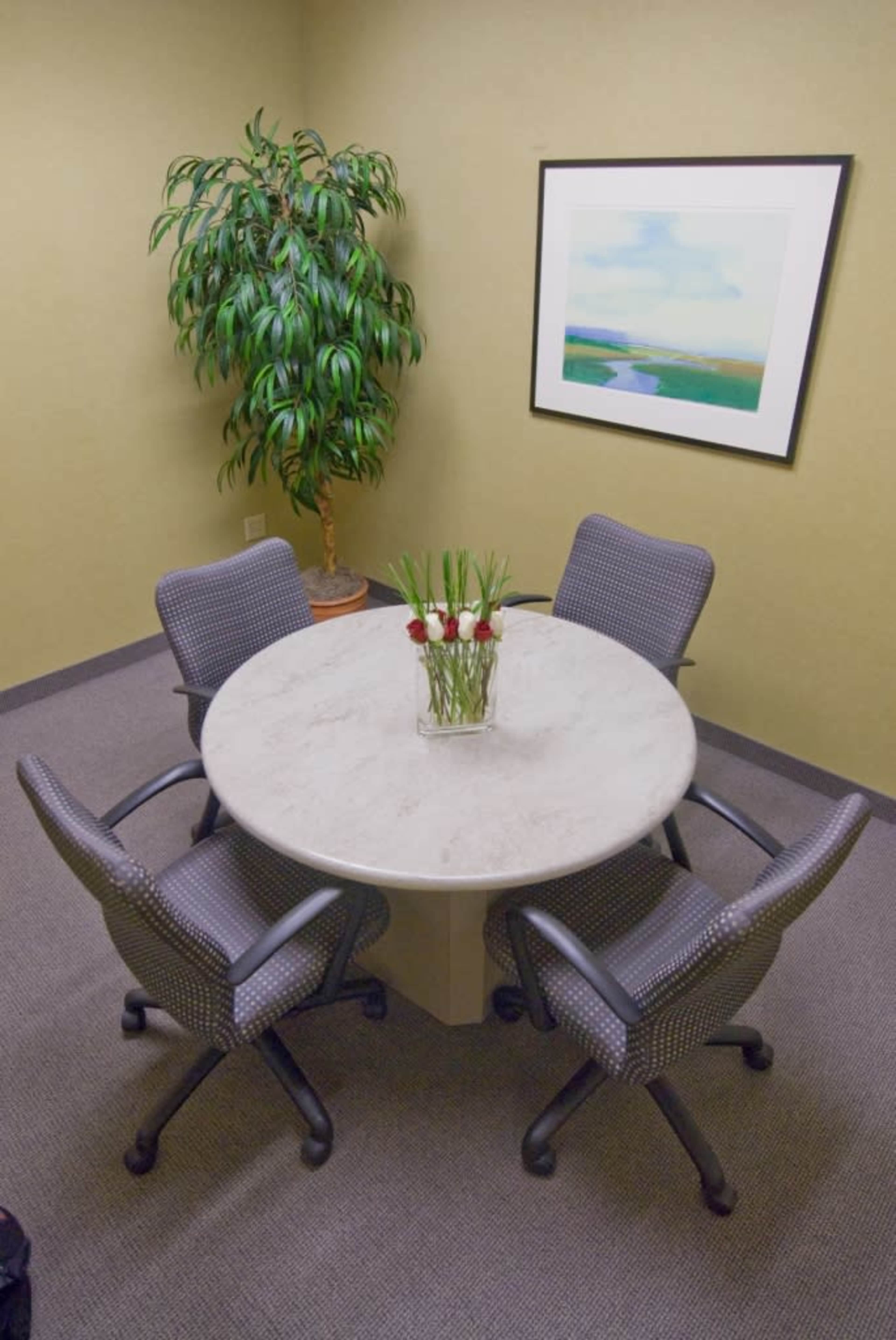 A round table with four chairs is set in a small meeting room, featuring a potted plant and a colorful framed artwork on the wall.