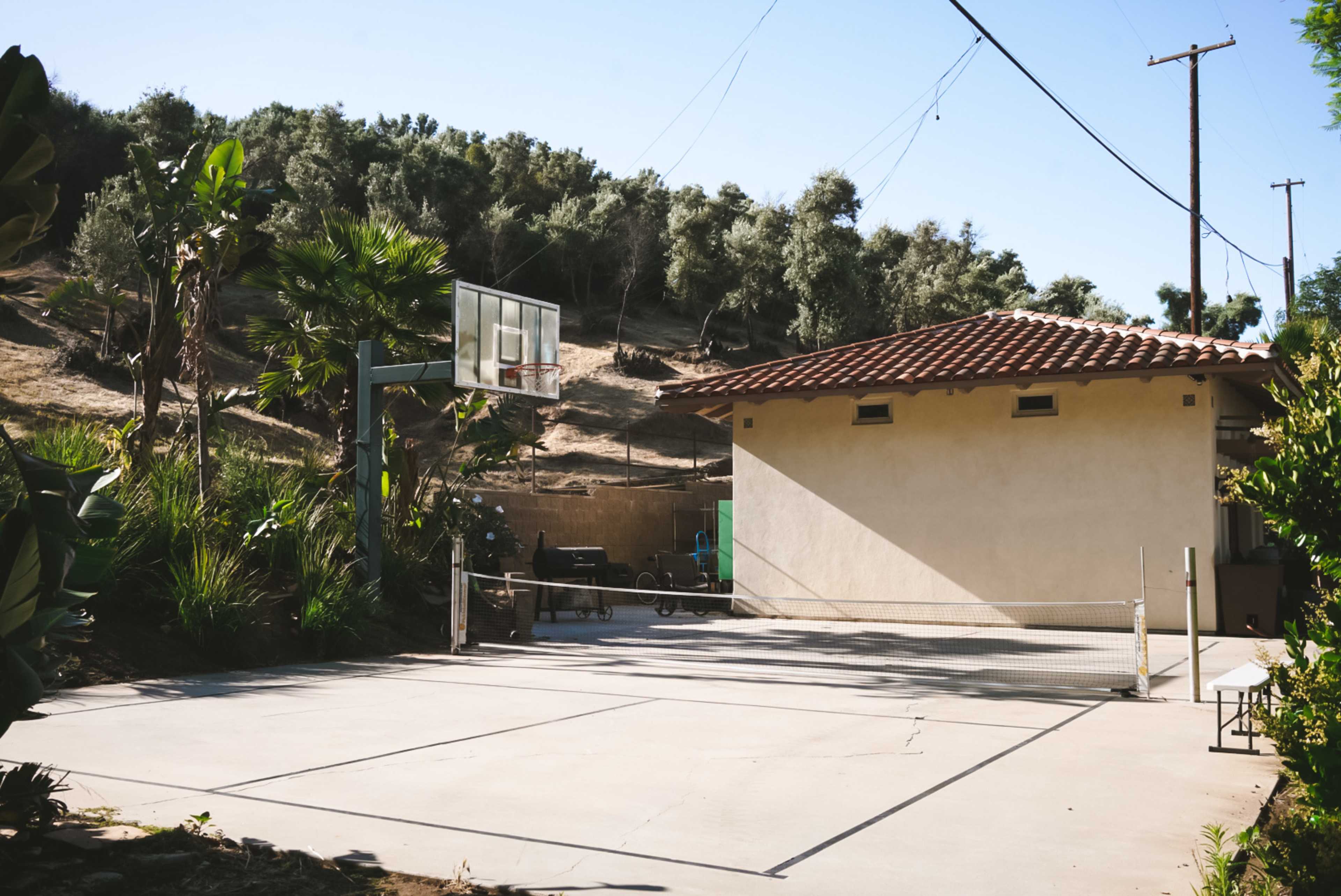 A concrete basketball court next to a light-colored building with a tiled roof, surrounded by greenery and hills in the background.