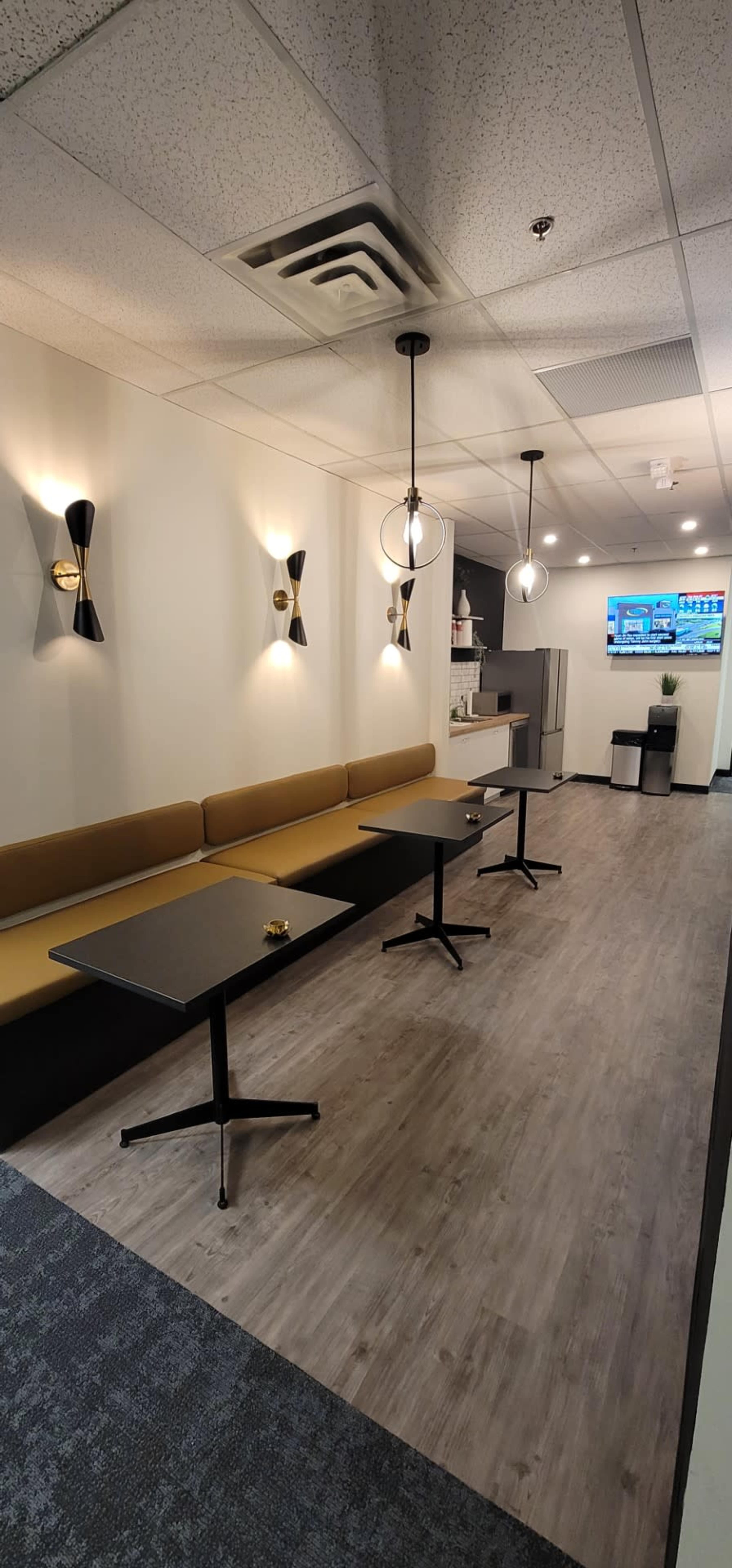 The image shows a modern lounge area with a long beige bench, two black tables, and pendant lights, leading to a kitchenette and a television display in the background.