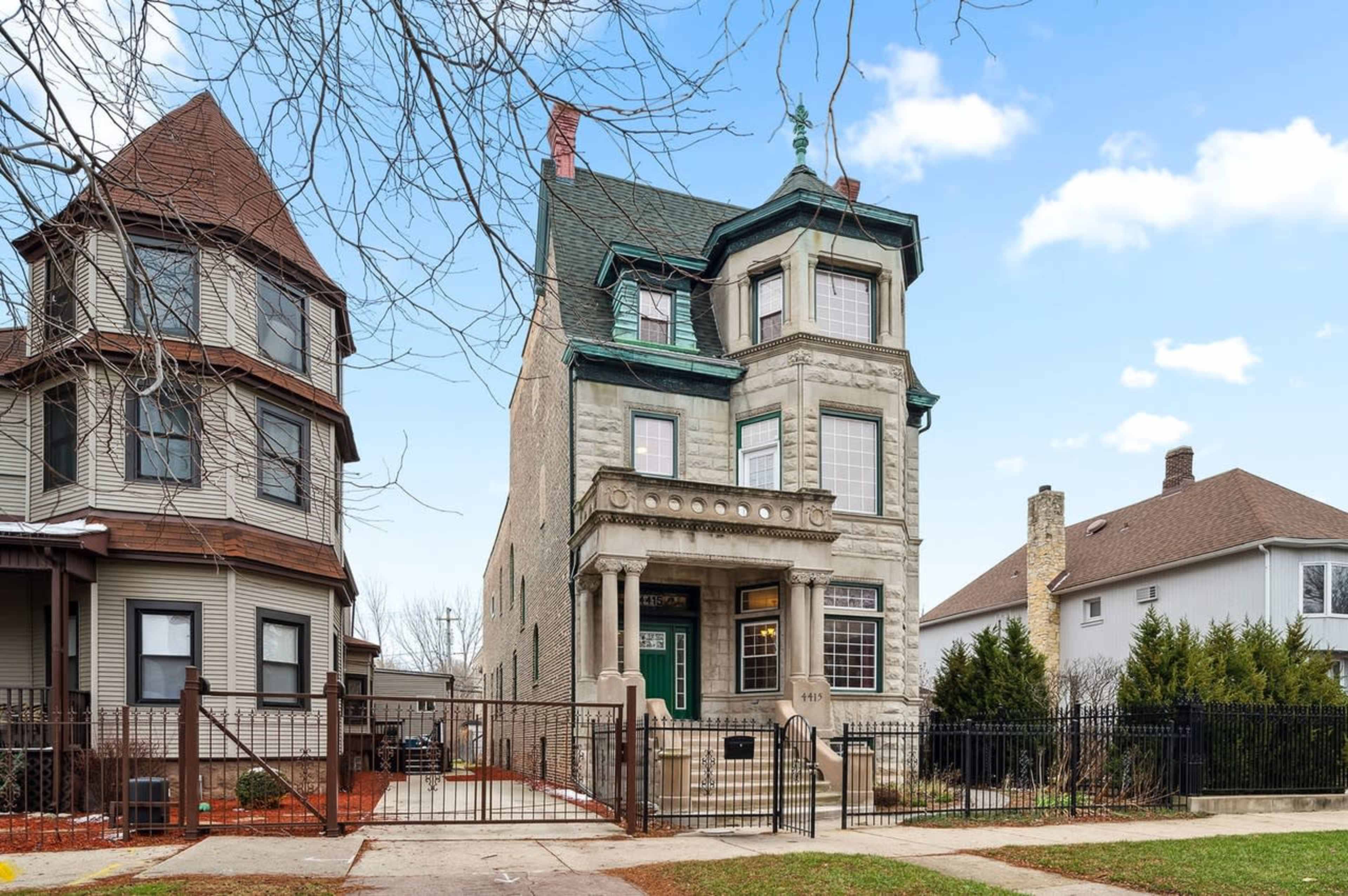 A large, three-story brick house with a green roof and decorative elements stands between two other residential buildings.