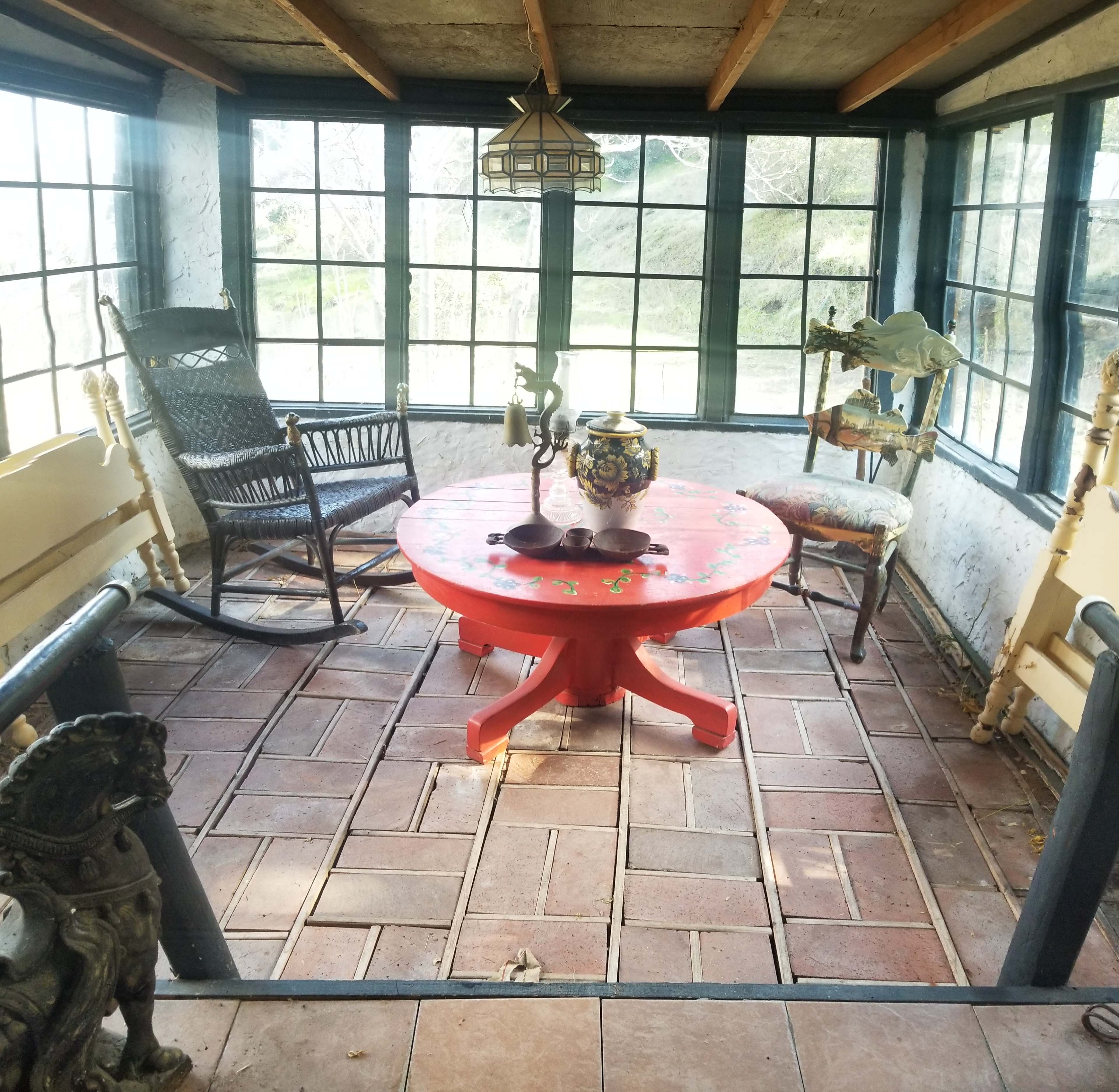 A sunlit room with large windows, featuring a red circular table, several chairs, and decorative items like a vase and a rocking chair.