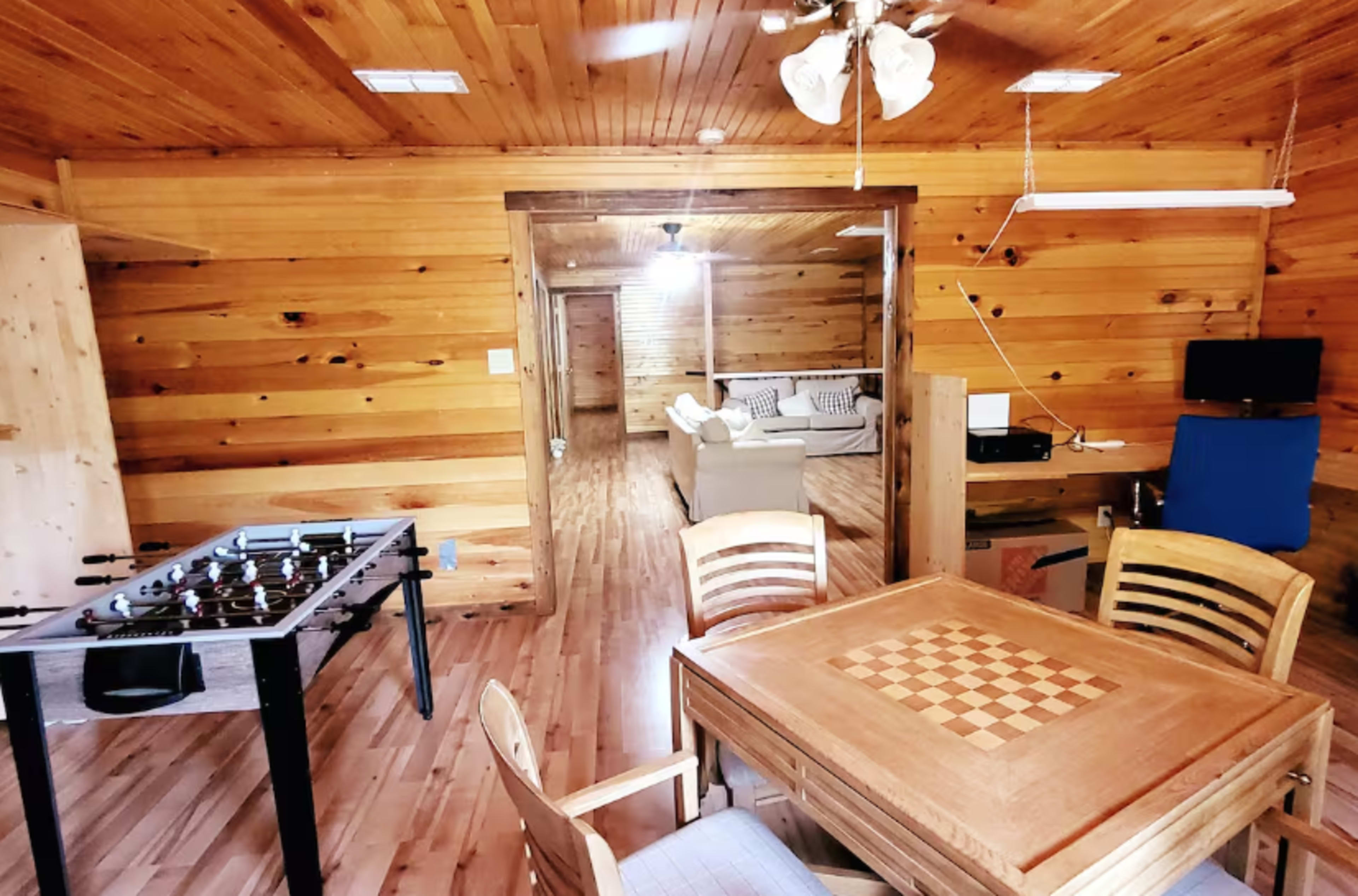 The image shows a cozy wooden room with a foosball table, a chess table, and a living area visible in the background.