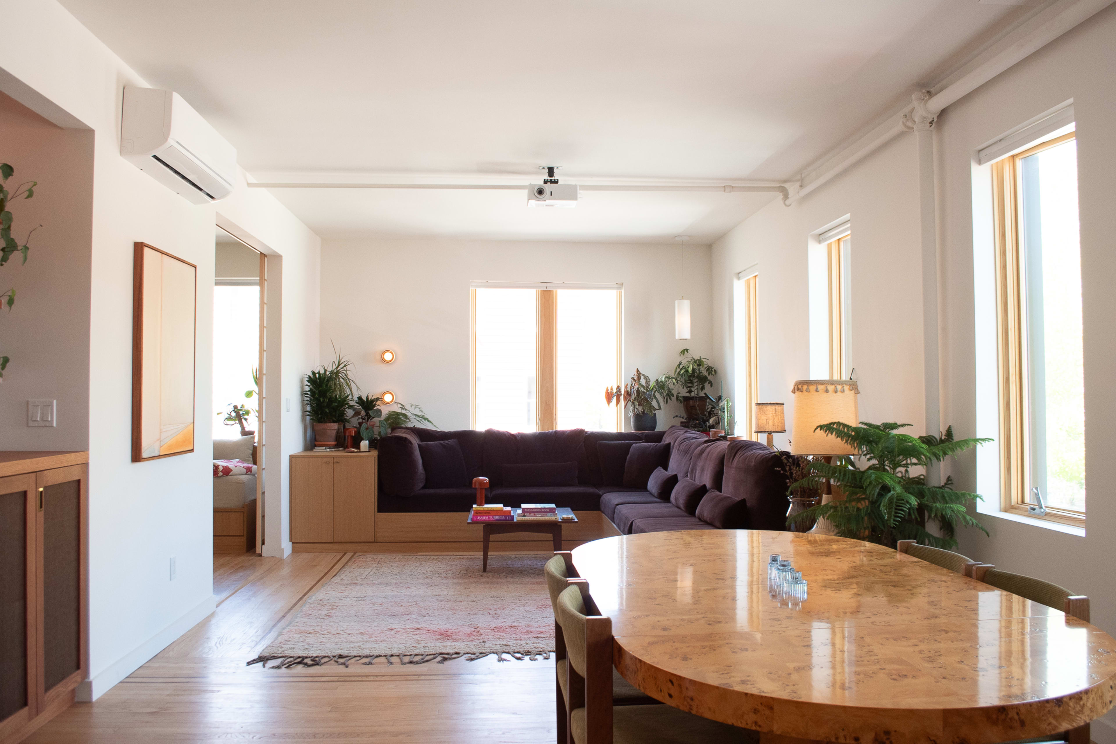 A modern living space features a sunlit area with a sectional sofa, a coffee table, and a dining table surrounded by chairs, along with plants and large windows.