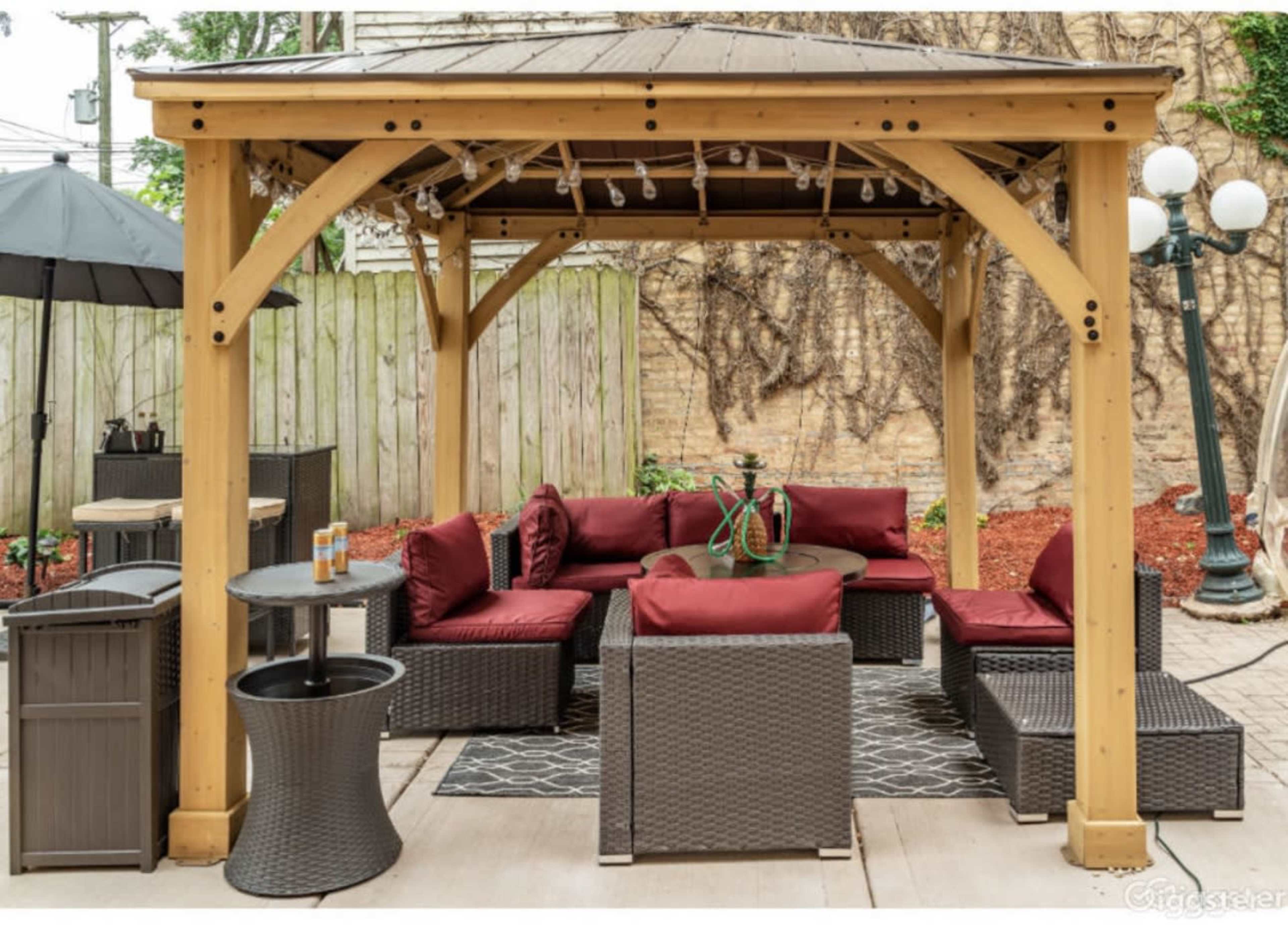 A wooden gazebo with a seating area consisting of dark wicker furniture and red cushions, surrounded by a landscaped patio.