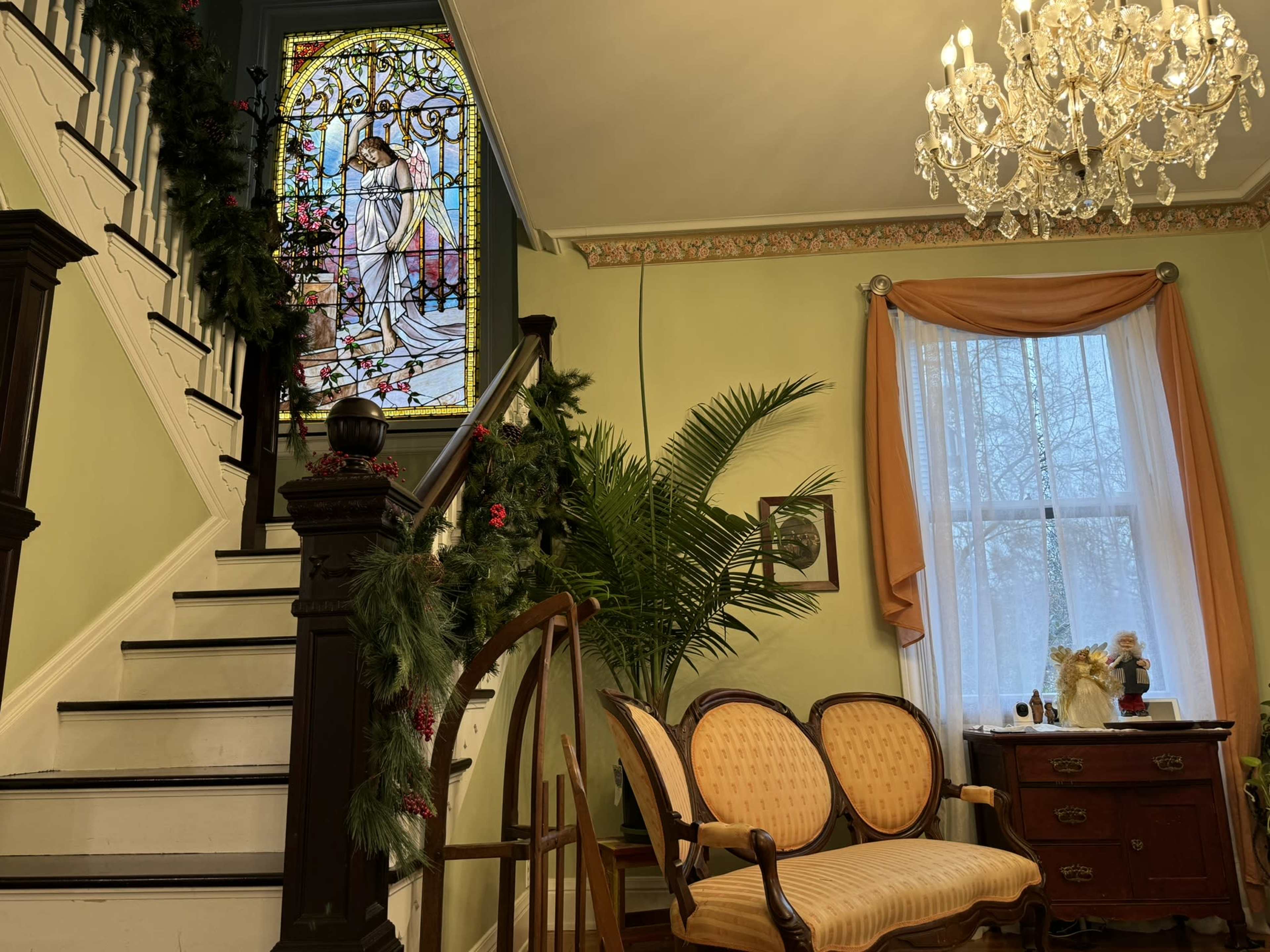 A staircase with a stained glass window, a vintage sofa, and a wooden desk in a room decorated with festive greenery and a chandelier.
