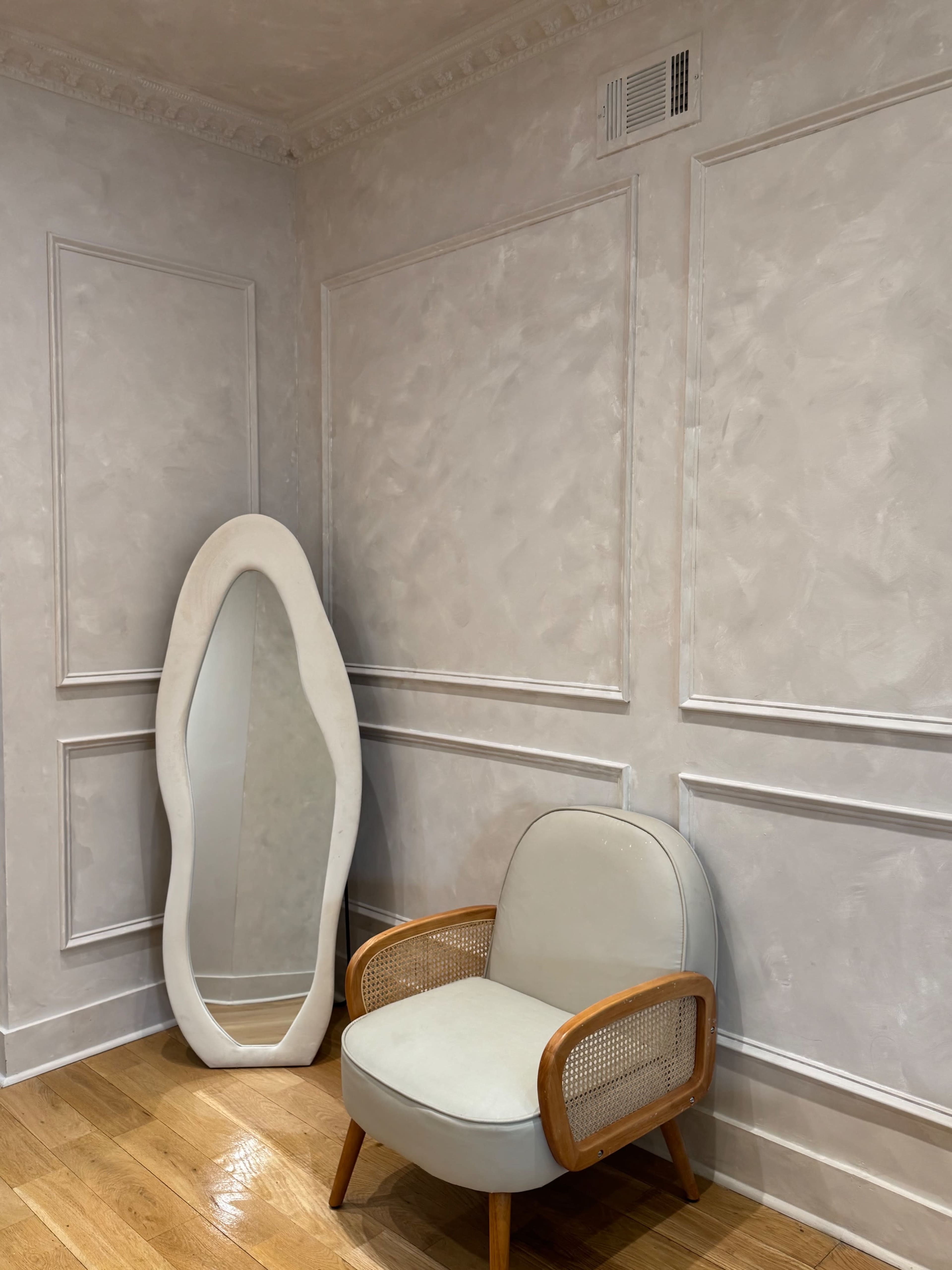 A light-gray room features a modern chair with wooden accents beside a uniquely shaped mirror mounted on the wall.
