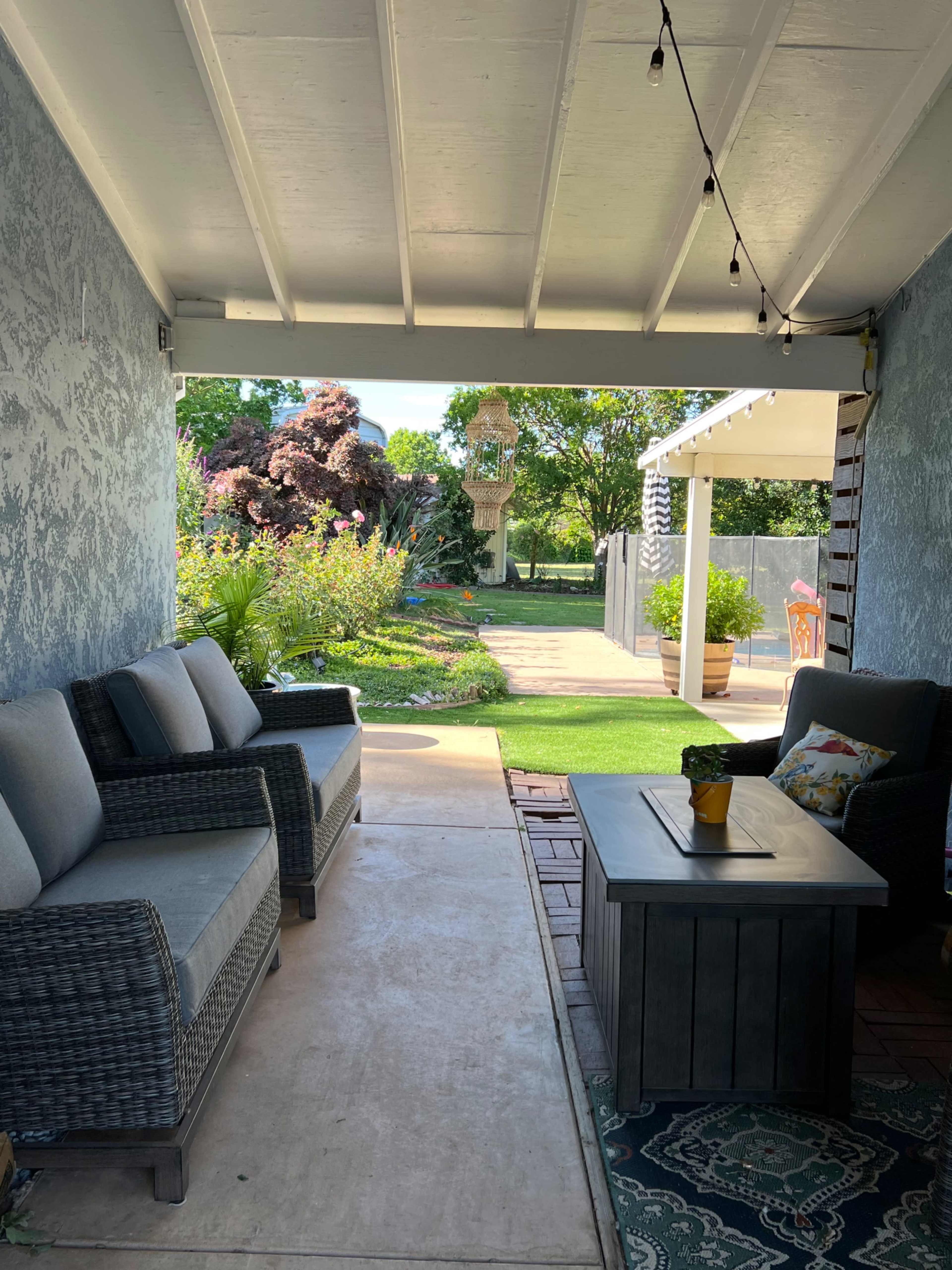 The image shows a covered outdoor seating area with wicker furniture, flanked by a well-maintained garden and a pathway leading to a patio.