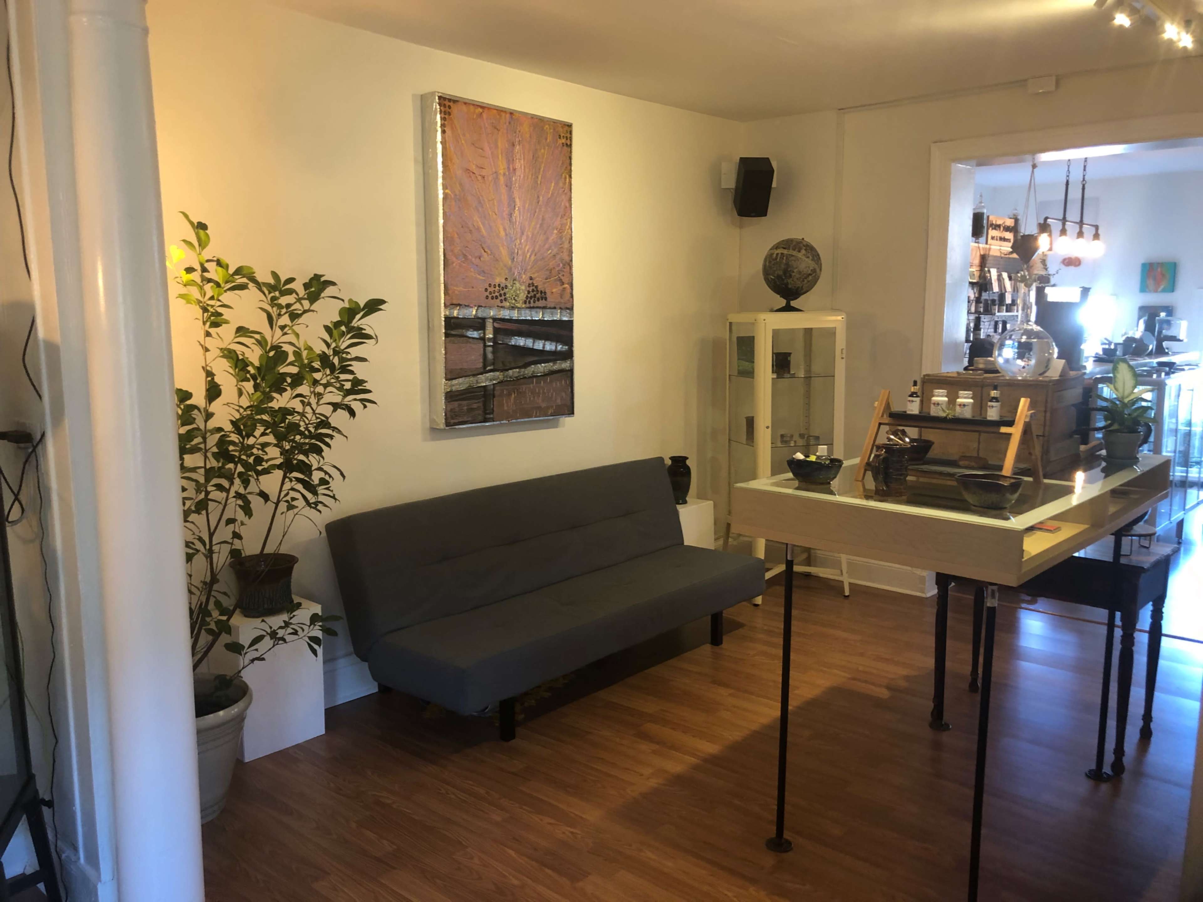 A cozy interior space featuring a gray sofa, a small table with items on it, and decorative plants, along with a large artwork on the wall.