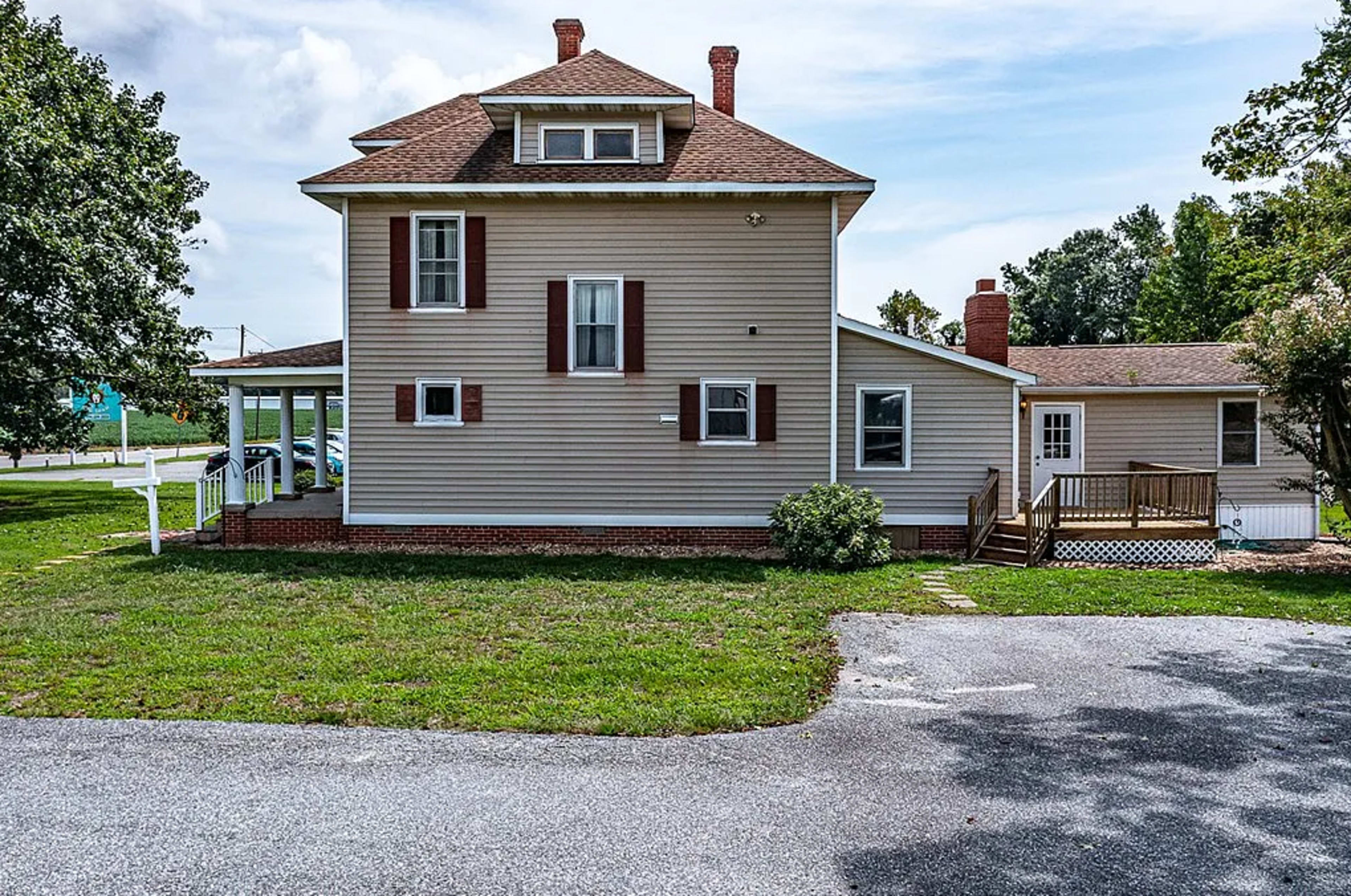 The image shows a two-story house with a beige exterior, red shutters, and a porch, situated on a grassy area with a paved driveway.