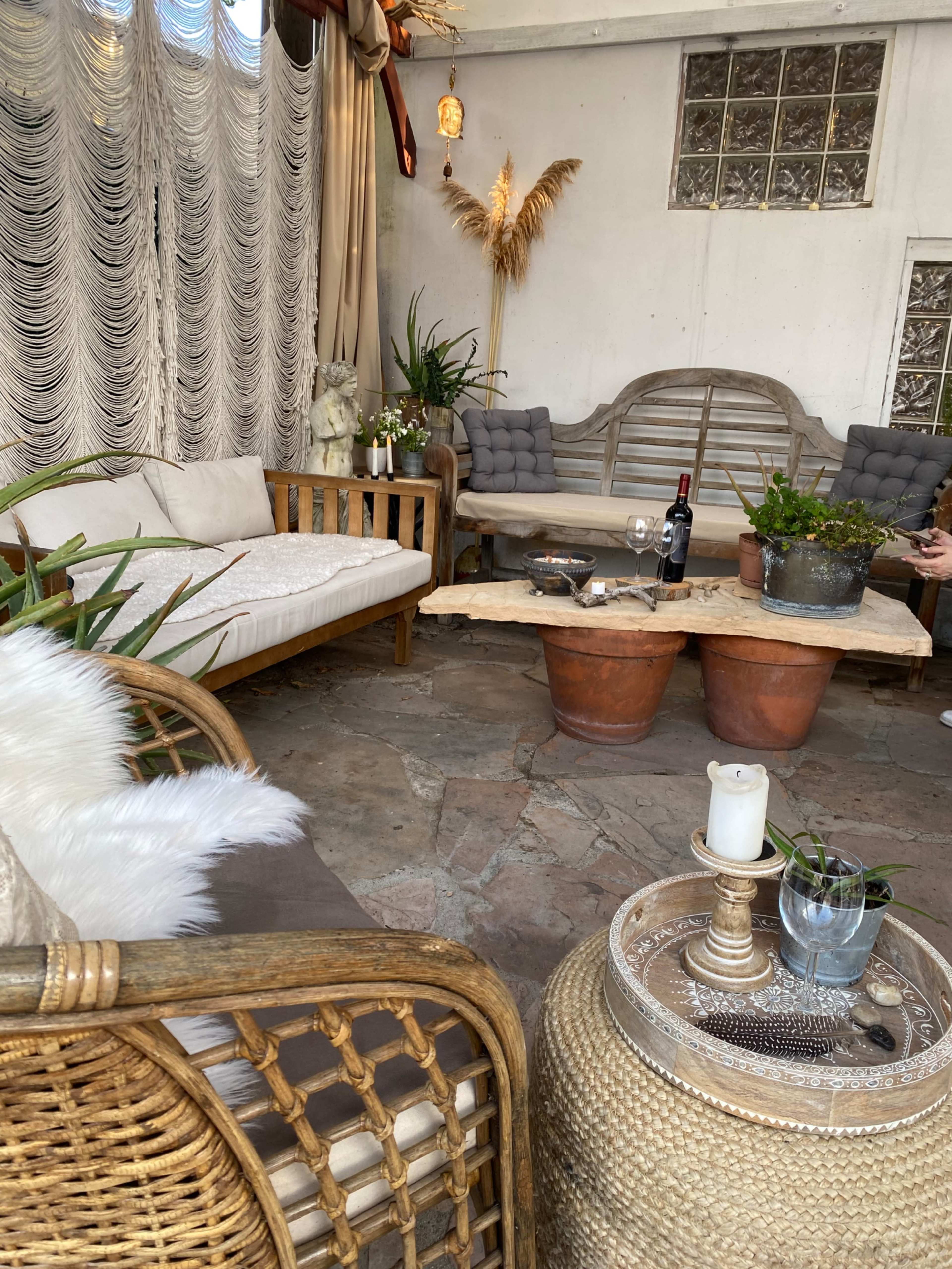 A cozy sitting area with rattan furniture, a wooden coffee table, potted plants, and a bottle of wine on a stone floor.