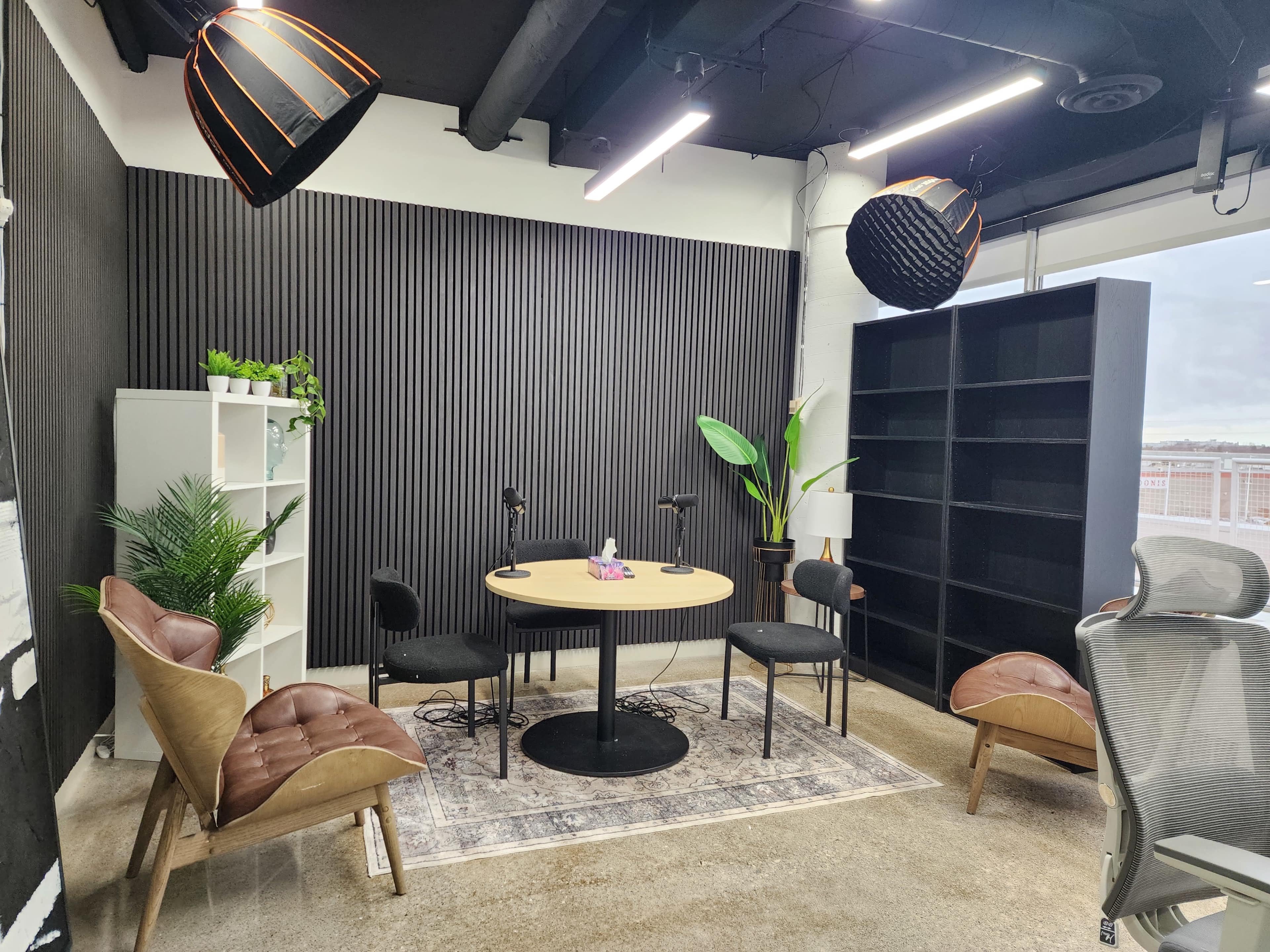 The image shows a modern workspace with a round table at the center, surrounded by chairs, and decorated with plants and shelves against a textured wall.