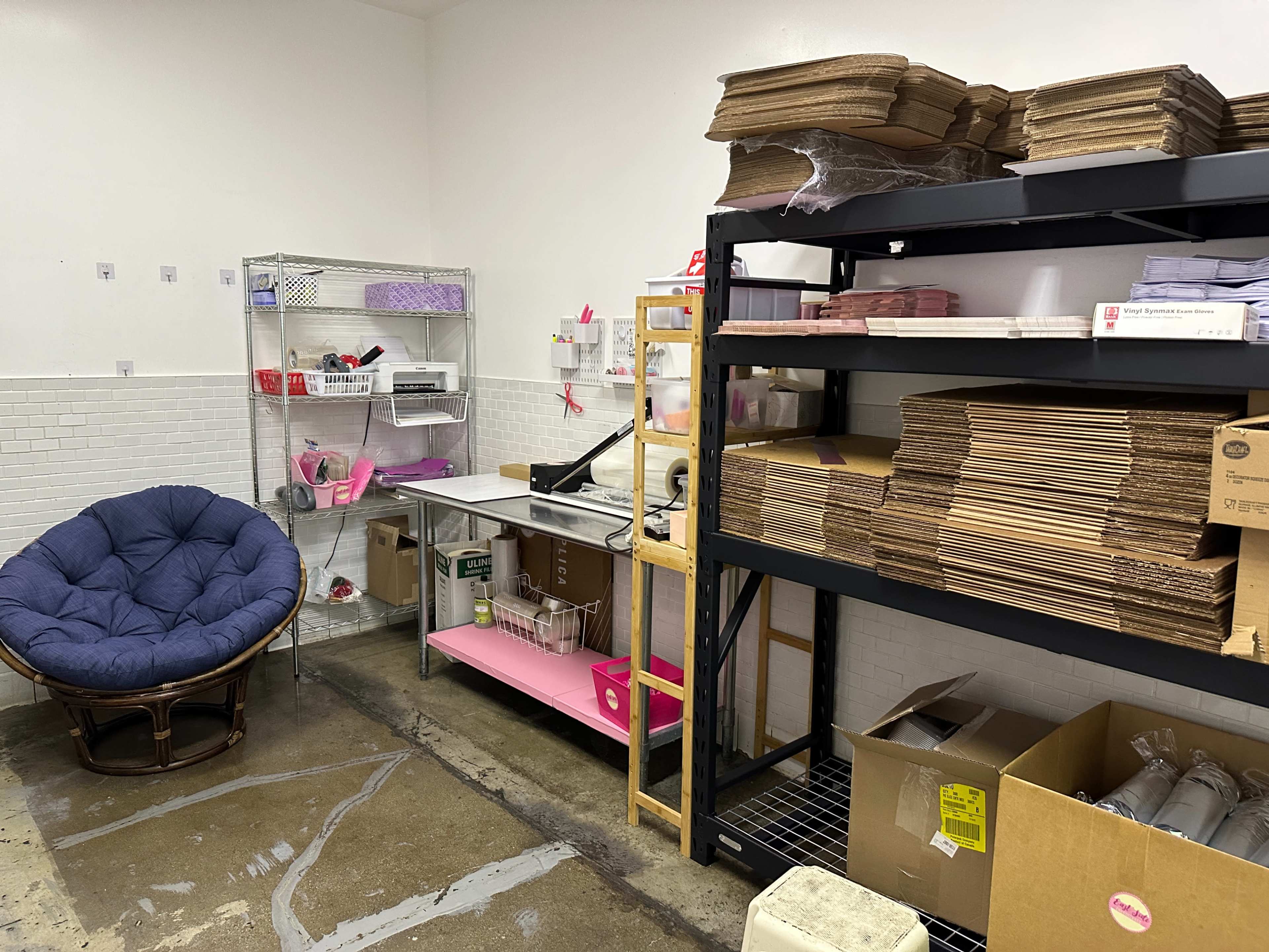 A storage room with a large blue chair, shelves filled with cardboard boxes, and a work table equipped with various supplies.