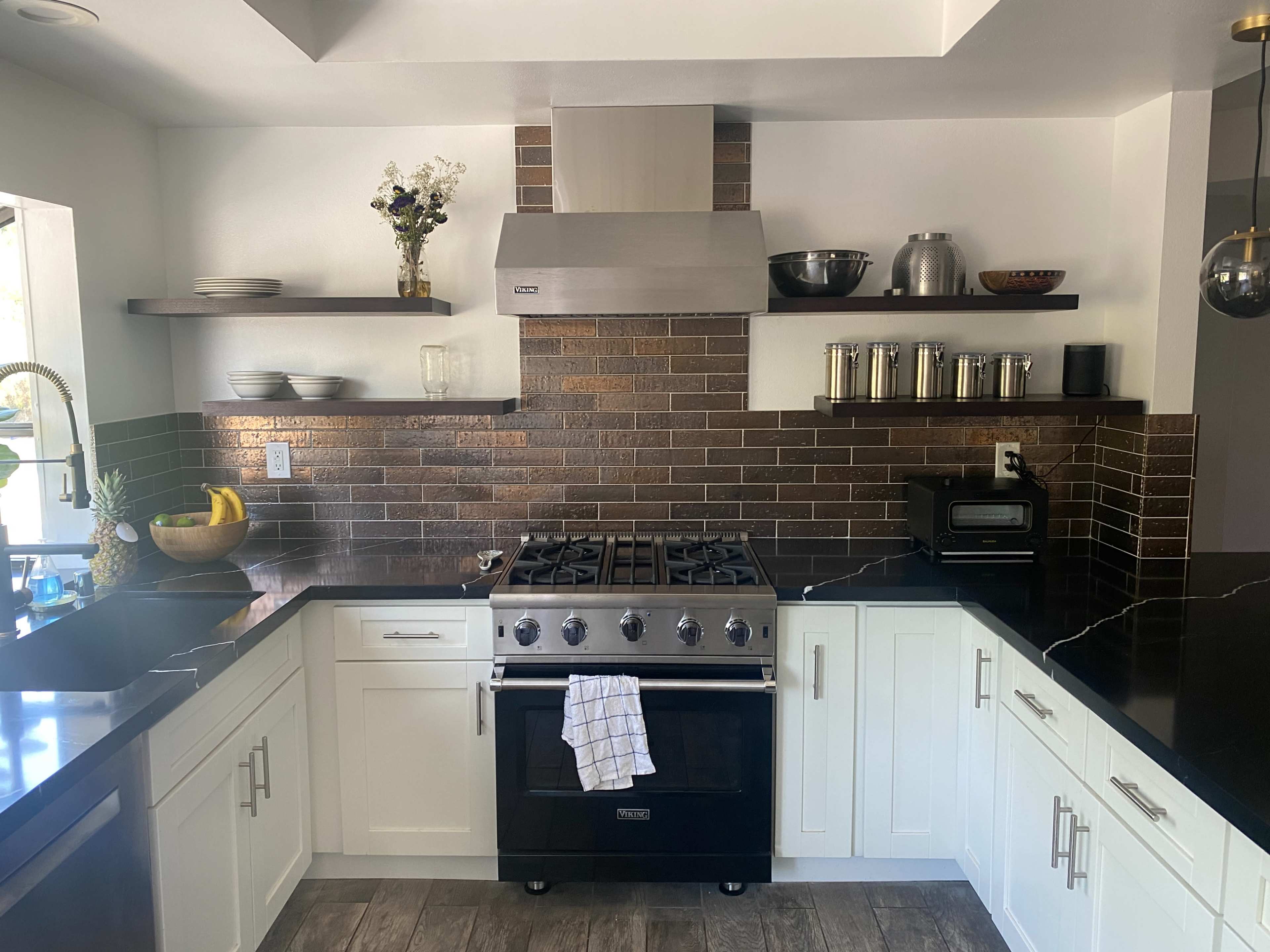 A modern kitchen features a stainless steel gas range, white cabinets, dark countertops, and open shelves with decorative items and cooking utensils.