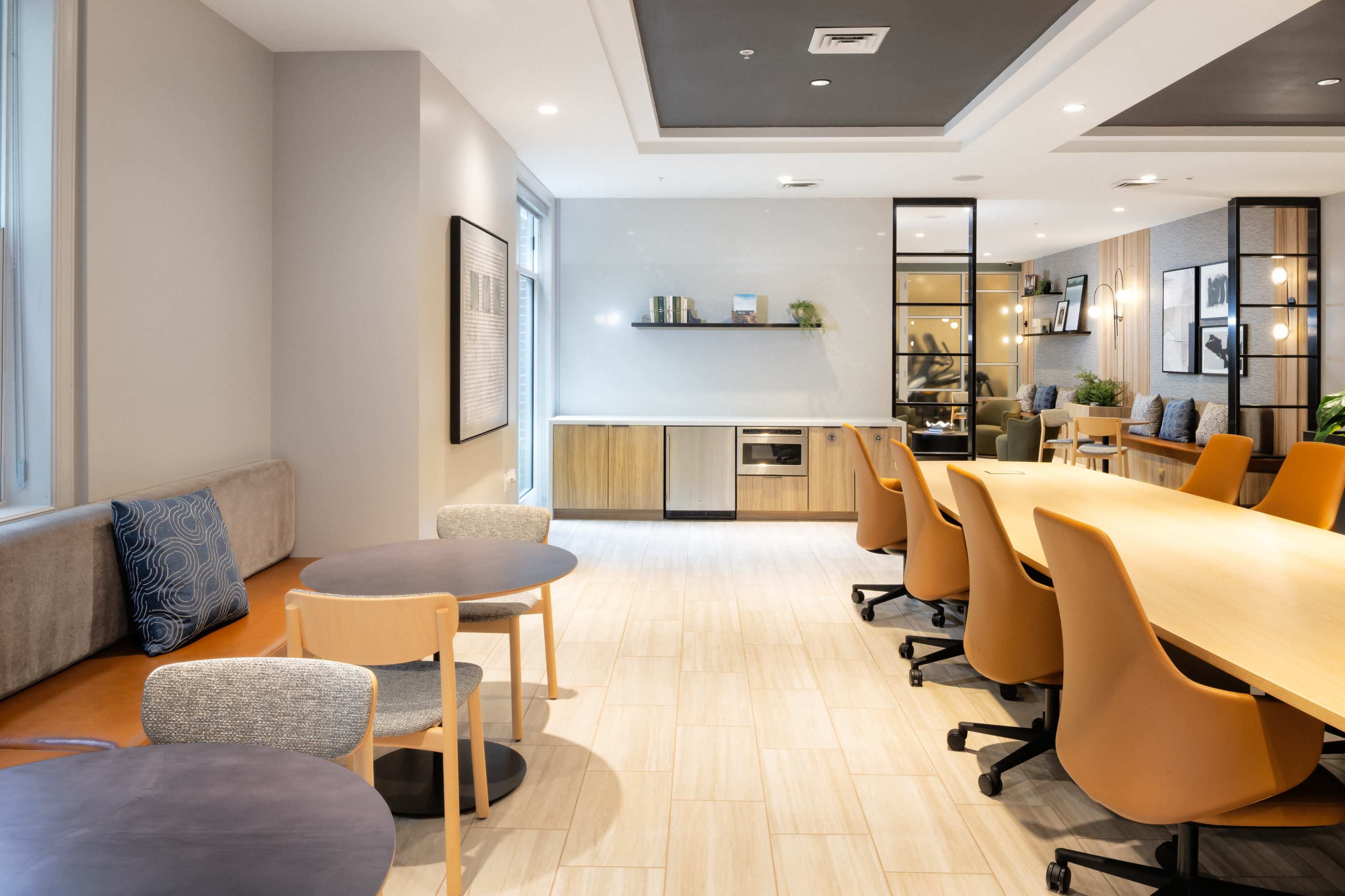 The image shows a modern interior space with a mix of seating options, including tables and chairs, along with a kitchenette area and large windows.