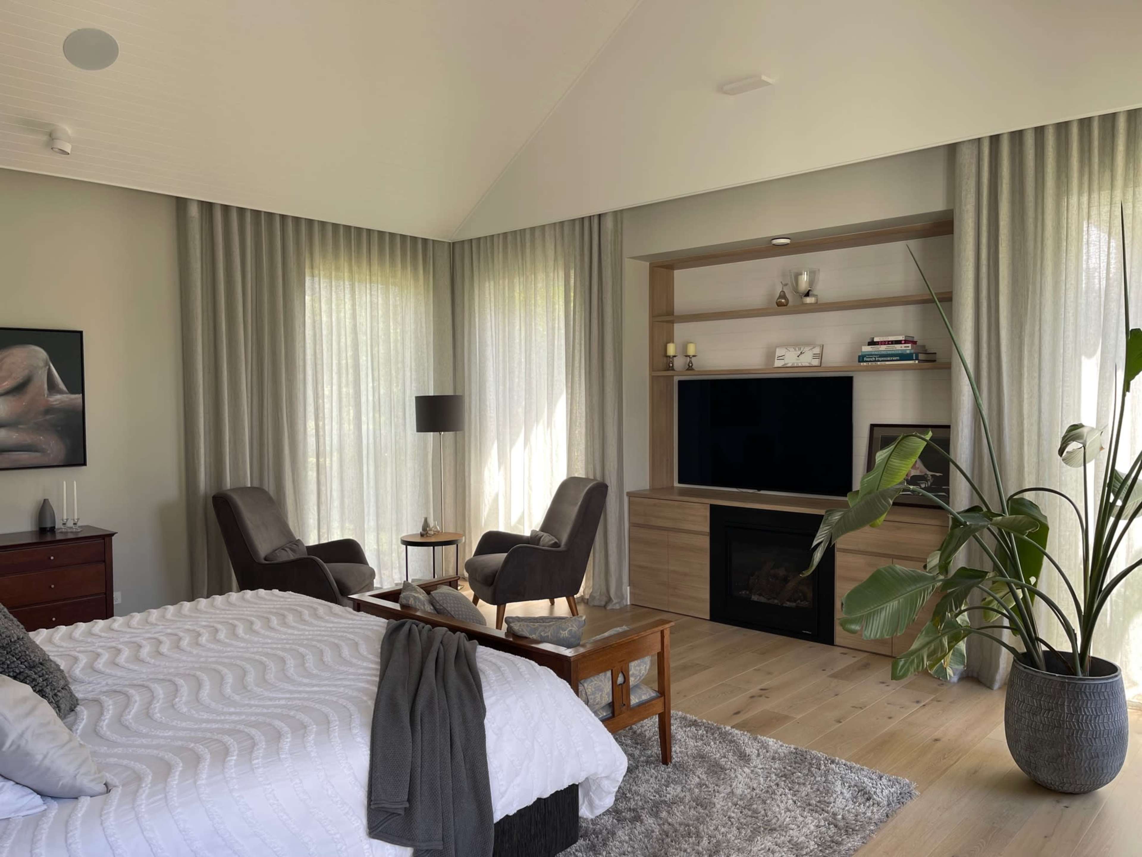 A modern bedroom features a bed with a textured white bedspread, two armchairs beside a television cabinet, and large windows covered with sheer curtains.