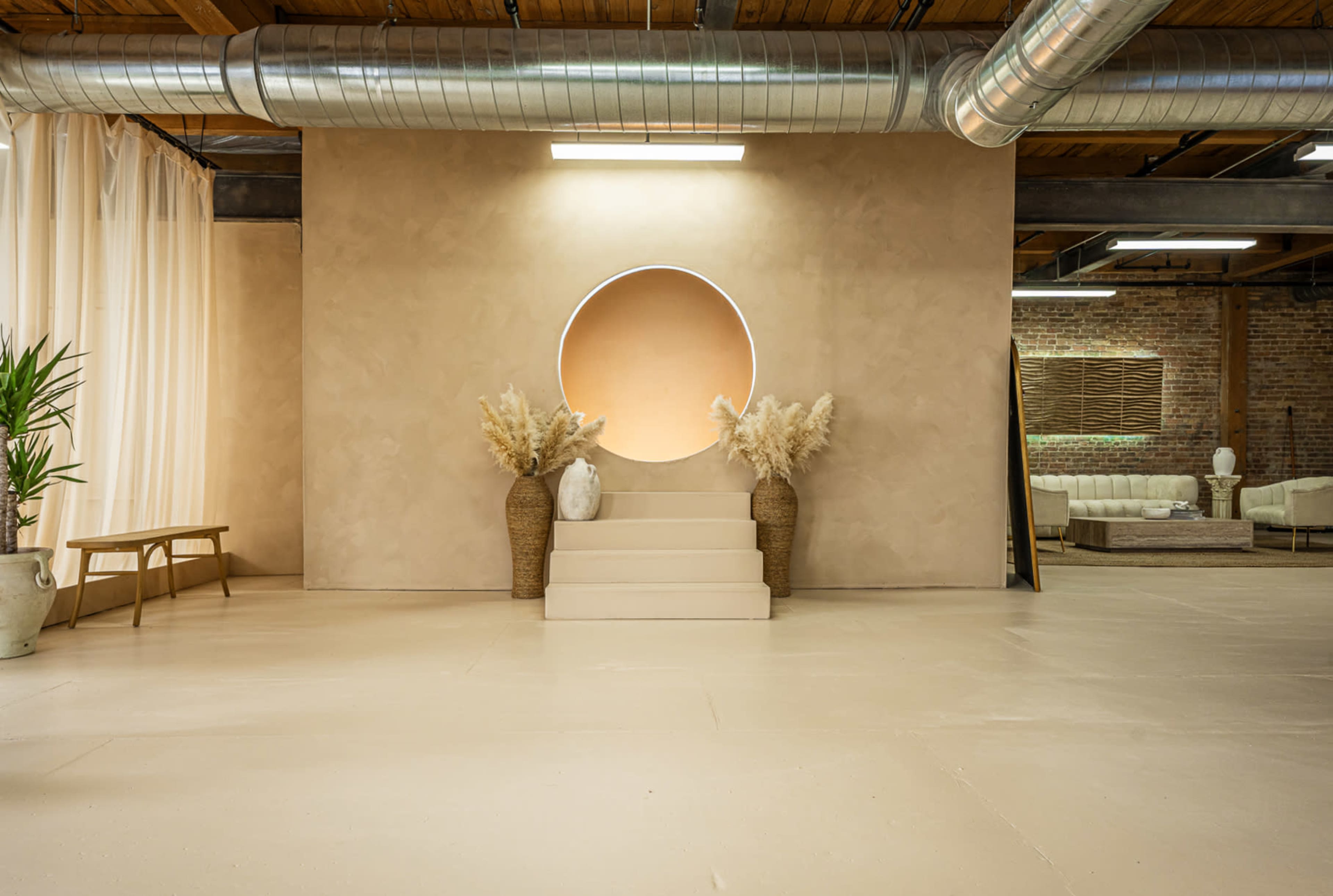 A minimalist studio space features a circular backdrop with warm tones, flanked by decorative plants and minimalist furniture.